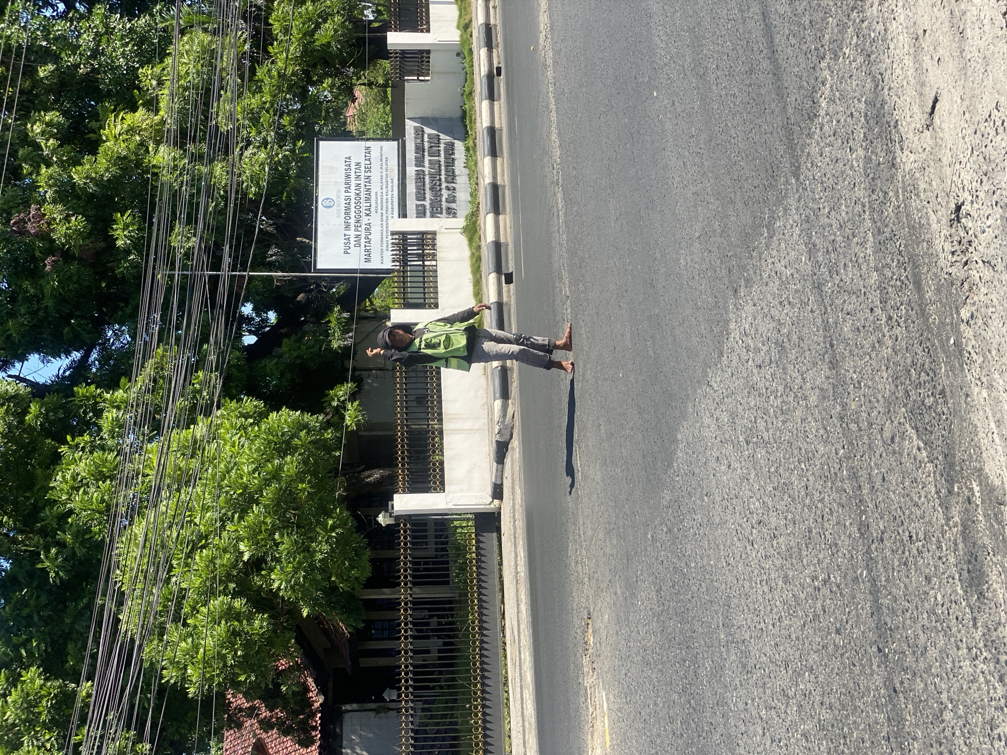 A single individual, wearing a high-visibility green vest and grey pants, stands in the middle of a paved road. The individual is barefoot and appears to be signaling with their left hand, which is raised. A large tree with lush green foliage dominates the background, with power lines strung across it. To the right of the tree, a white sign is visible, bearing text in Indonesian. The sign appears to be an information center, with the words "PUSAT INFORMASI PARIWISATA DAN PENGGOSOKAN INTAN MARTAPURA - KALIMANTAN SELATAN" visible. Behind the sign and the tree, a low-rise building with a tiled roof and a black metal fence is partially visible. The road surface is asphalt with visible tire marks and some debris. The scene appears to be in a tropical or subtropical climate, given the dense foliage and bright sunlight. The location is identified as Sungai Paring, Indonesia.