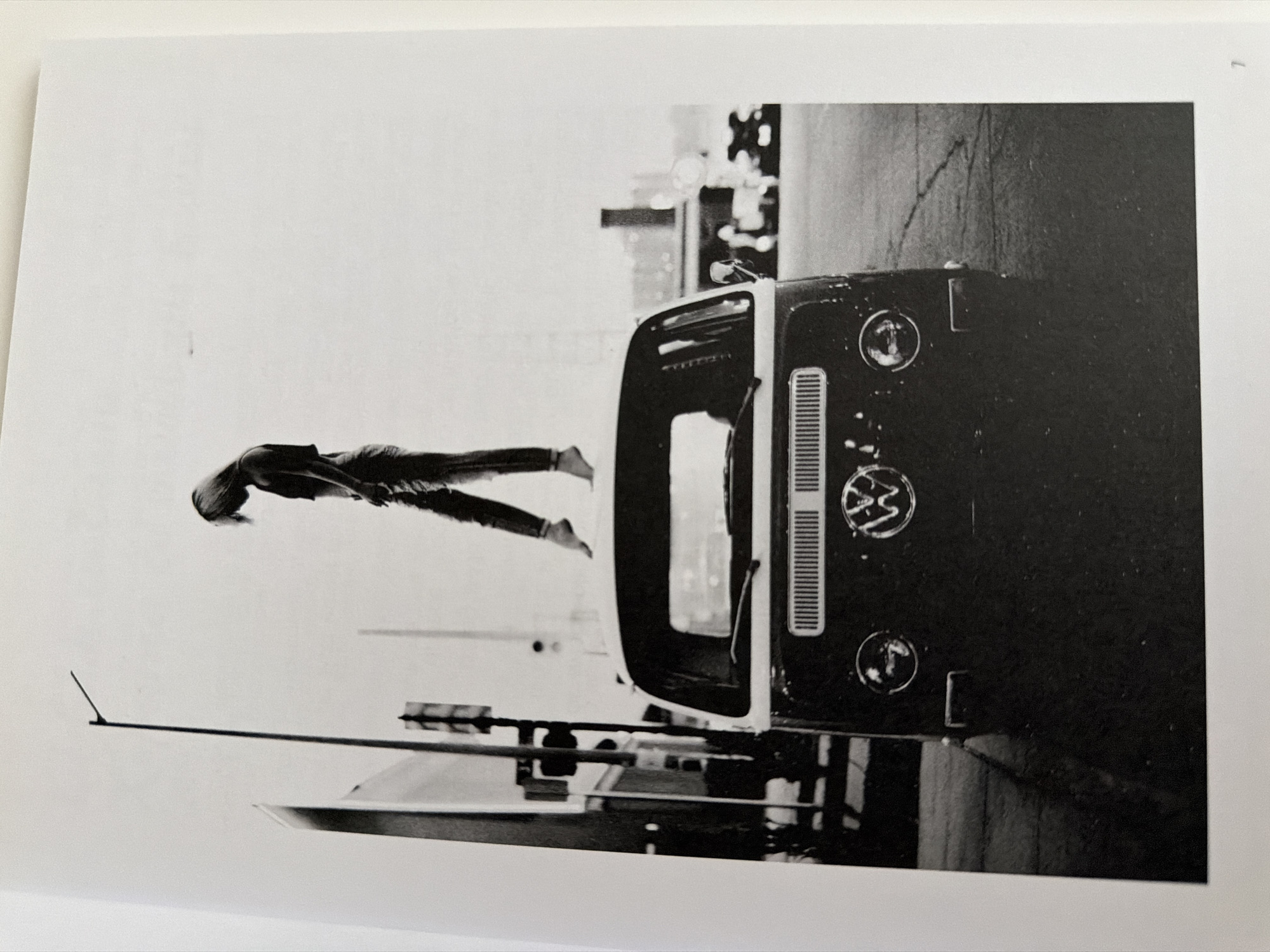 A black and white photograph shows a woman standing on the roof of a vintage Volkswagen bus. The bus is dark, and the woman is wearing light-colored jeans. She is facing away from the camera, with her head bowed. The background is a bright, overcast sky with a street lamp and some buildings visible. The scene appears to be on a street in a city, but the specific location in Pazardzhik, Bulgaria, cannot be determined from the image. The composition is dynamic, with the woman's pose creating a sense of both vulnerability and defiance. The overall mood is somewhat melancholic, perhaps suggesting a moment of introspection or escape.