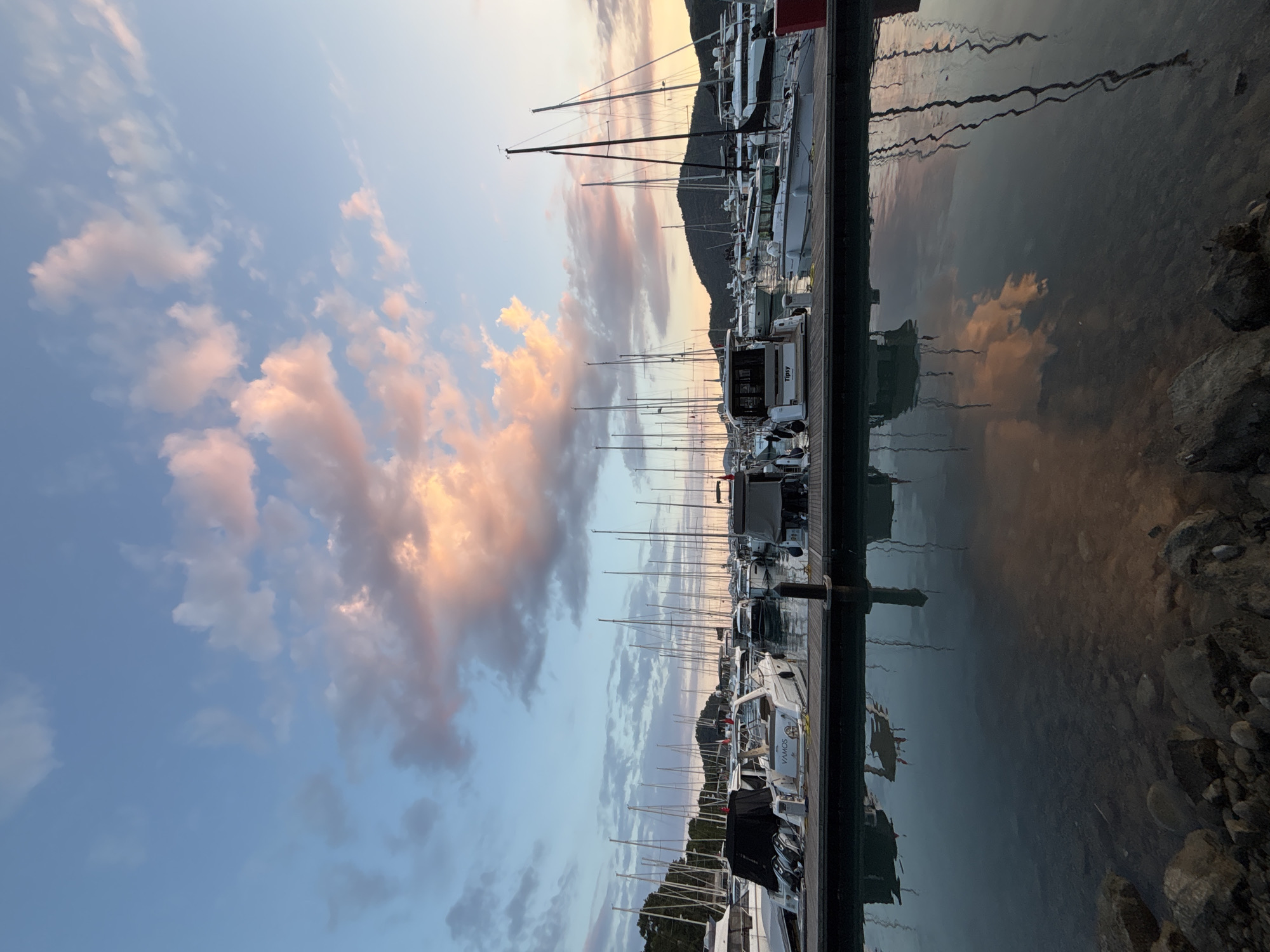 This image depicts a serene marina in Fethiye, Turkey, during what appears to be sunset or twilight. The sky is a gradient of soft blues and pinks, with scattered clouds illuminated by the setting sun. Numerous sailboats and motorboats are docked along a wooden pier, their masts forming a striking vertical pattern against the sky. The calm water of the marina perfectly reflects the boats and the colorful sky, creating a symmetrical and tranquil scene. Rocky shorelines are visible in the foreground. The overall atmosphere is peaceful and picturesque.