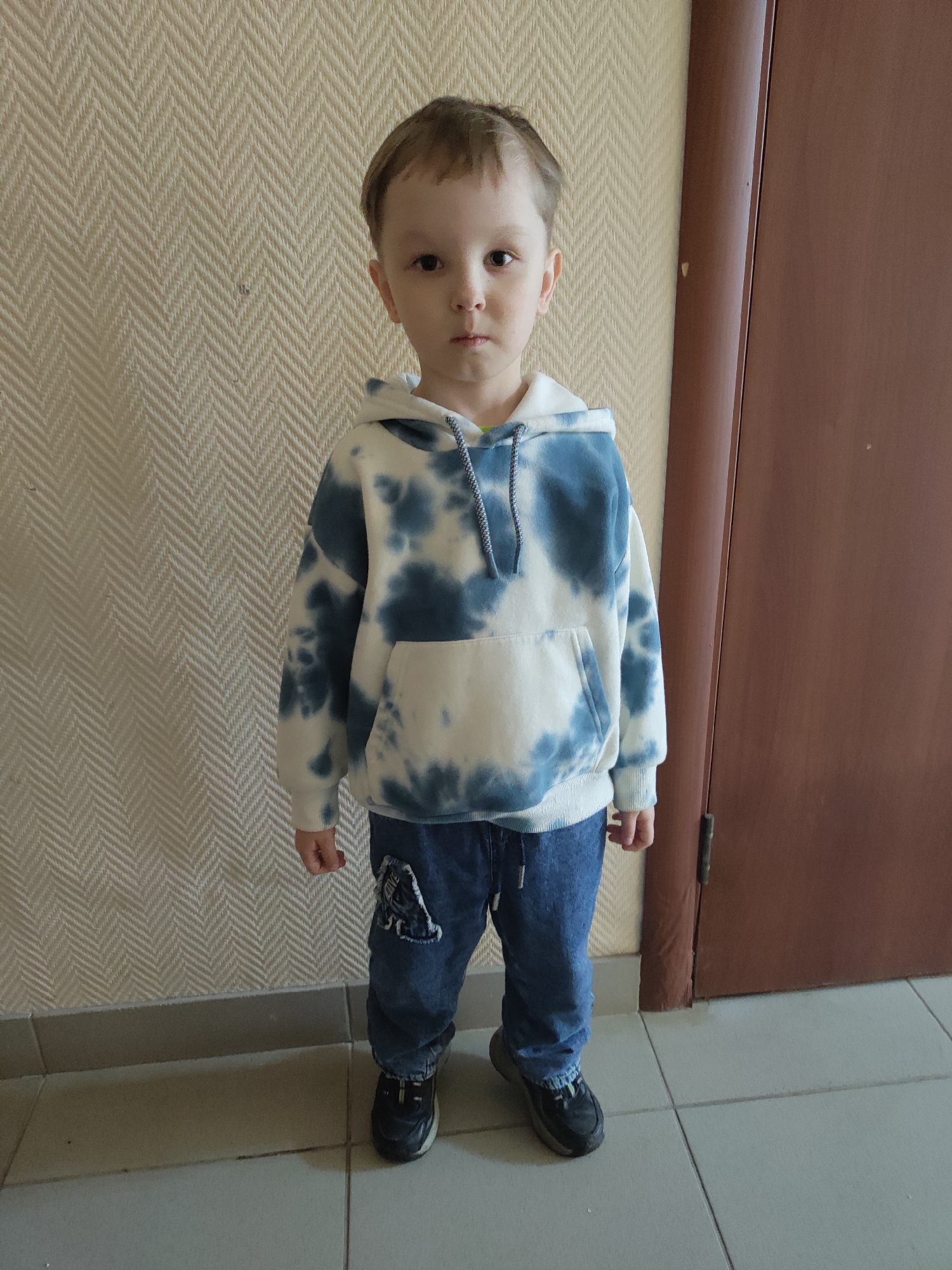 A young child, likely a boy, is depicted in a full-body portrait, standing centrally and facing the camera with a neutral expression. He has short, light brown hair. He is dressed in a white and blue tie-dye hooded sweatshirt with a front kangaroo pocket and a grey drawstring. He wears blue denim jeans, which have a distressed patch on the upper left leg (viewer's right) with some partially obscured text. His footwear consists of dark-colored sneakers with light-colored soles.
The child is positioned against a light beige or cream-colored wall covered in wallpaper featuring a distinct herringbone or chevron pattern. To the right of the child (viewer's left), a plain, dark brown wooden door or doorframe is visible, extending from the top to the bottom of the frame. The floor is covered with light grey square tiles, separated by darker grout lines, and a darker grey baseboard runs along the bottom of the wall.
The scene takes place indoors, likely in a hallway or room, under even lighting. There is no specific activity occurring other than the child posing for the photograph. The location context is Chaikovsky, Russia.