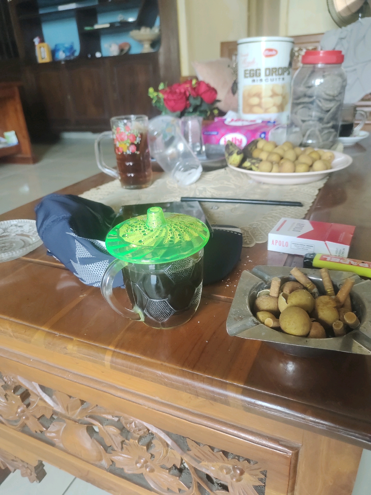 The image captures a close-up view of a wooden coffee table in what appears to be a domestic setting in Rejowinangun, Indonesia. The table is cluttered with various items. In the foreground, there is a glass of dark liquid, possibly coffee or tea, with a bright green plastic lid. To the right of the glass, an ashtray is filled with cigarette butts and what look like small, round fruits, possibly longans. Next to the ashtray is a pack of "APOLO" cigarettes and a yellow lighter.
Further back on the table, there is a plate piled with more of the small, round fruits. Behind that, a glass jar with a red lid contains what appear to be dried noodles or similar food items. A tin of "Egg Drops Biscuits" is also visible. In the background, a bouquet of red flowers adds a touch of color. A dark blue cap lies near the glass of drink. The wooden furniture, including a carved cabinet in the background, suggests a traditional Indonesian style. The overall impression is of a casual, lived-in space where someone has been enjoying snacks and drinks.