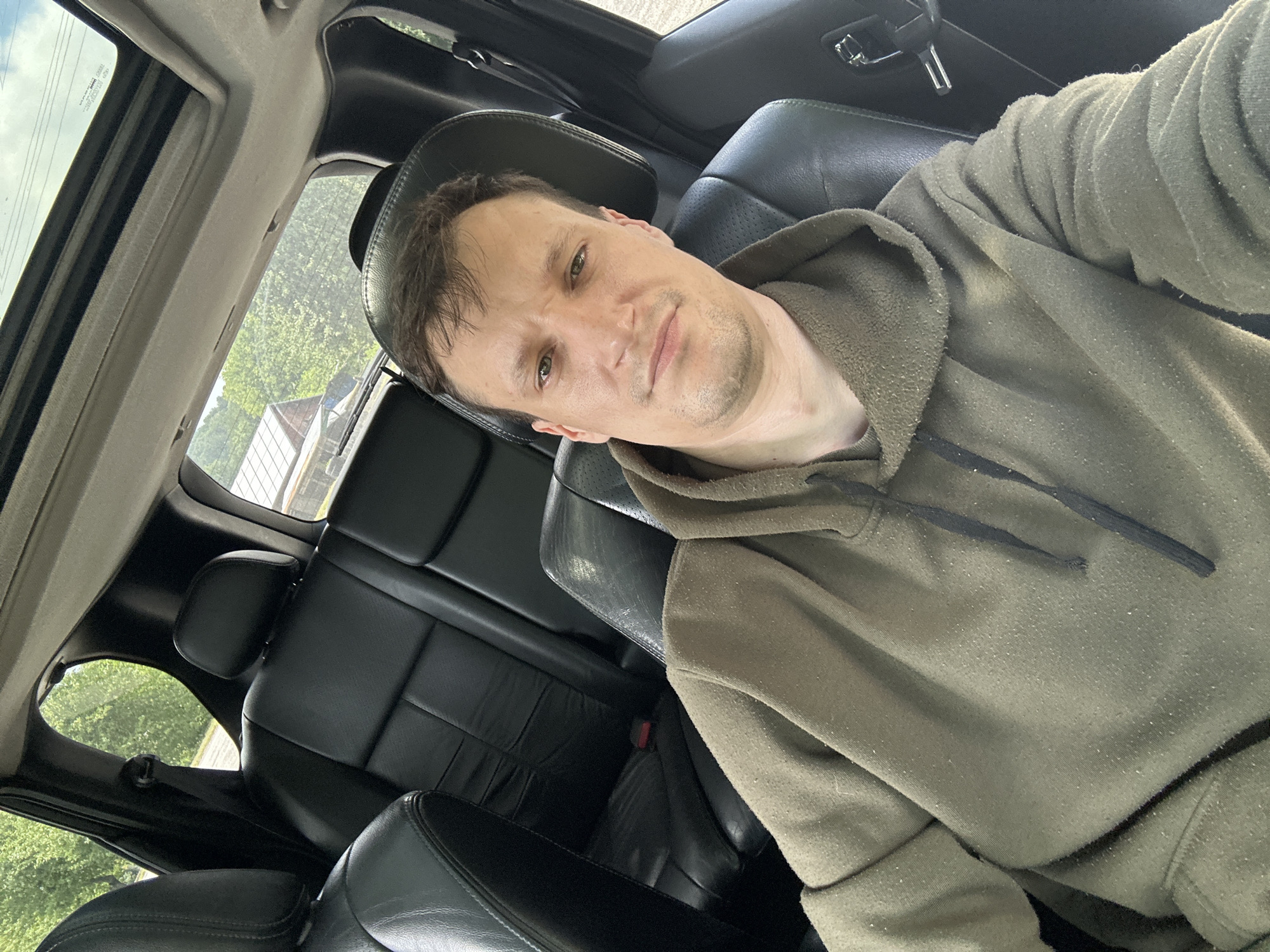 A man is pictured inside a car, taking a selfie. He is wearing a green hoodie and is seated in the driver's seat. The car's interior is visible, with black leather seats and a sunroof. Outside the car, trees and a building can be seen through the windows, suggesting a rural or suburban setting. The image does not provide specific information about the location in Makiv, Ukraine.