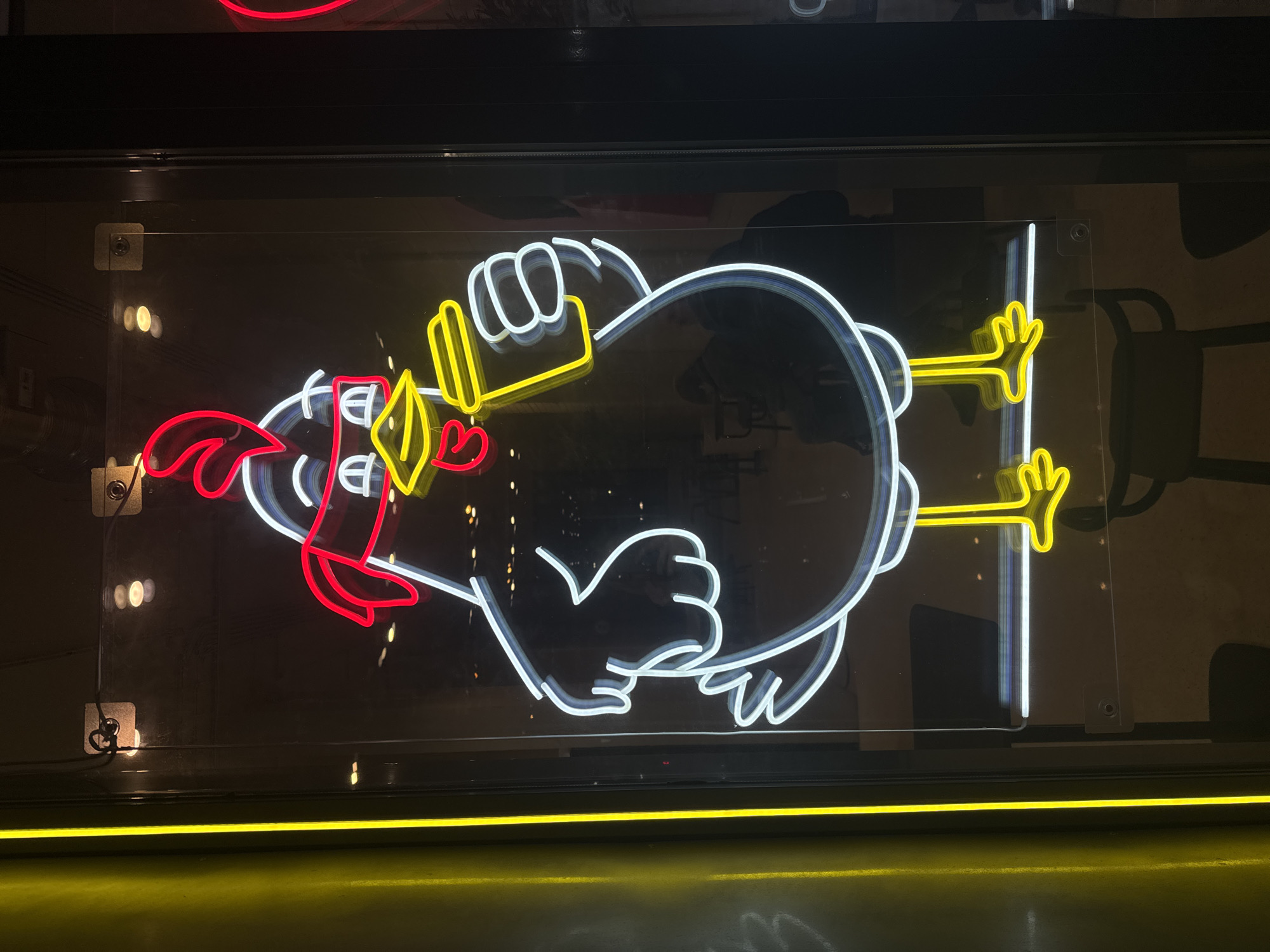 The image shows a neon sign of a chicken displayed in a storefront window. The sign features a white outline of a chicken with red accents on the comb, wattle, and a small heart. The chicken has yellow legs and feet, and is depicted holding a yellow dumbbell. The sign is mounted on a clear acrylic sheet with visible mounting hardware. The background is dark, showing reflections and hints of the interior space beyond the window. The bottom of the image is illuminated by a yellow light source, possibly a strip light. The storefront is likely located in a city, potentially Saint Petersburg, Russia, though there is no explicit visual confirmation of the location.