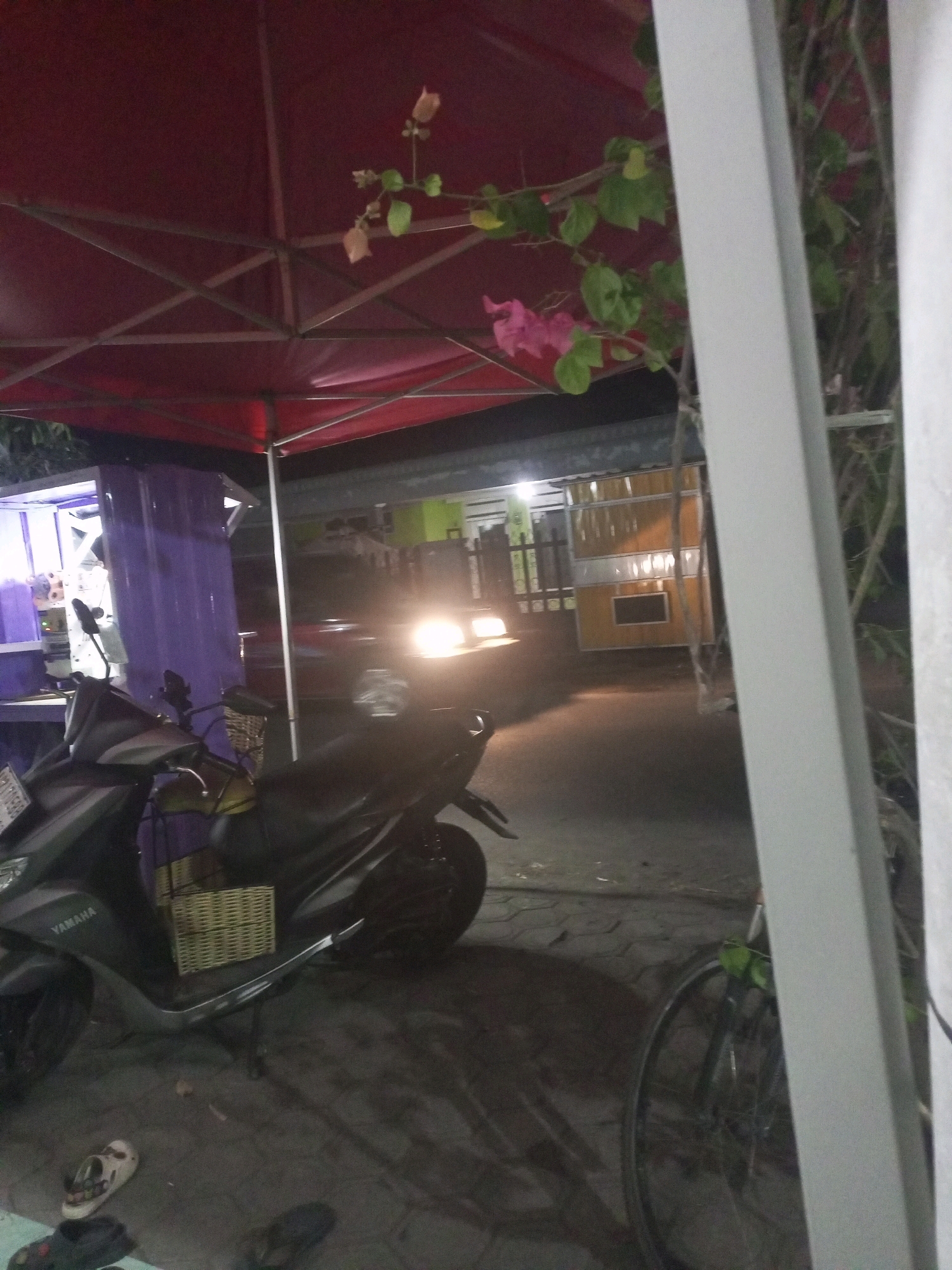 The image depicts an outdoor scene at night, likely in the city of Kediri, Indonesia, given the context. A red canopy, possibly part of a street stall or temporary structure, dominates the upper portion of the frame, supported by a metal frame. Bougainvillea vines with pink and light orange flowers are visible, cascading down from the canopy.
In the foreground, a black Yamaha scooter is parked. It has a wicker basket attached to its rear, and the "YAMAHA" logo is visible on the side panel. The scooter's headlight is off. A bicycle with a visible wheel and spokes is parked to the right of the scooter, partially obscured by a light grey vertical beam.
The ground appears to be paved with interlocking stones. A pair of sandals and other footwear are scattered on the ground near the scooter.
In the background, a street or pathway is visible, illuminated by artificial light. A red vehicle with its headlights on is moving or parked in the distance, creating a bright glow. Further in the background, there are structures with yellow and white panels, suggestive of a commercial area or residential buildings. A purple curtain or panel is visible on the left side of the frame, attached to a stall-like structure. The overall lighting suggests it is nighttime.