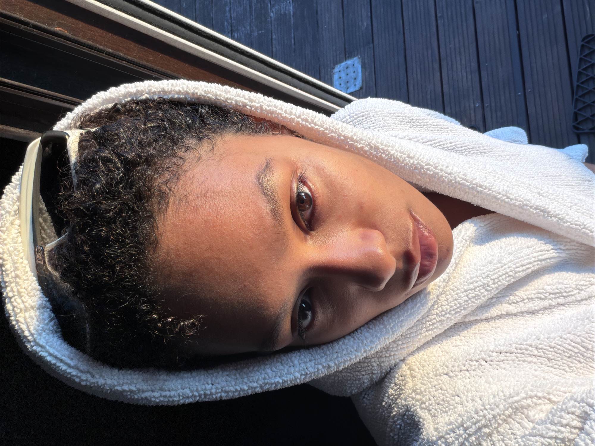 A young woman with curly dark hair and light brown eyes is shown from the chest up, looking directly at the camera. She is wearing a white bathrobe with the hood pulled up over her head, framing her face. Sunglasses are resting on top of the hood. The lighting suggests it is either early morning or late afternoon, with warm sunlight illuminating the right side of her face and the bathrobe, creating a strong contrast with the dark background on the left. She is positioned in what appears to be an outdoor setting, with a wooden deck visible in the background to the right, suggesting a balcony or patio. The overall atmosphere is serene and introspective. The image was taken on the terrace of the Château de Champs-sur-Marne.