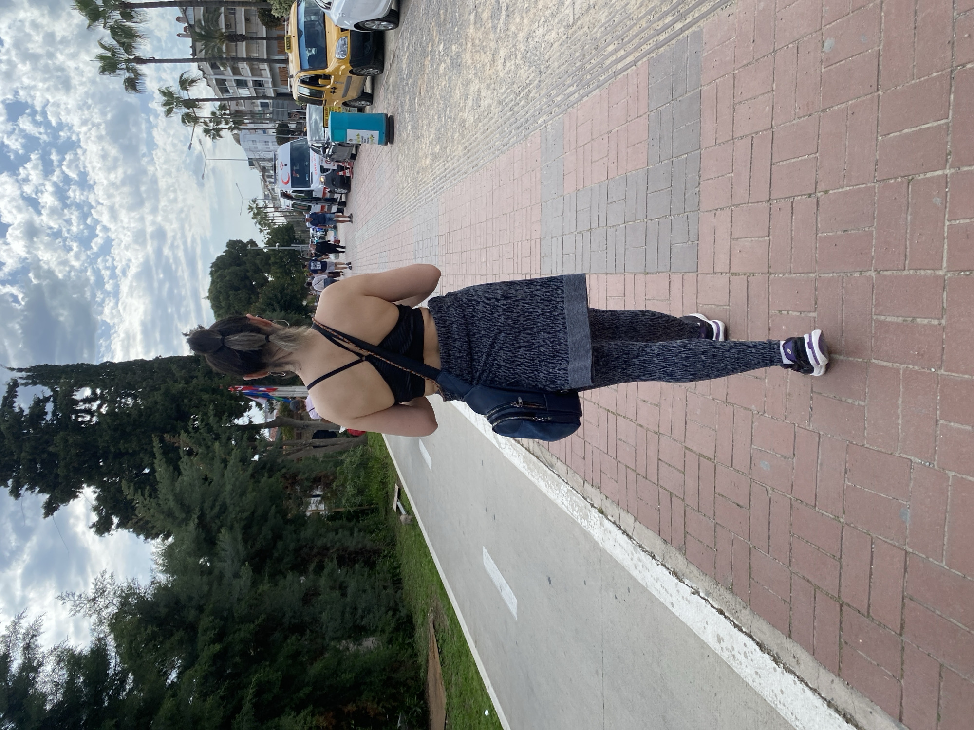 A woman is walking on a brick sidewalk alongside a bike path in Alanya, Turkey. She is wearing a black sports bra and patterned leggings with a dark blue crossbody bag. The sidewalk is next to a two-lane bike path with a white line separating the lanes. To the left of the bike path is a grassy area and trees. On the right of the sidewalk, a street with parked cars and pedestrians stretches into the distance. The weather appears partly cloudy, and palm trees line the street. Buildings are visible in the background.