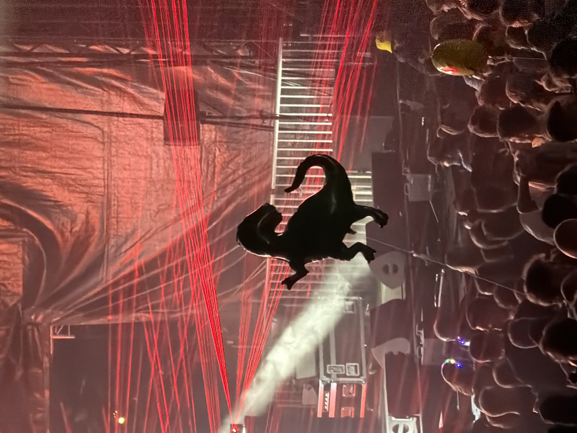 The image depicts a concert or event taking place at an indoor venue, possibly the Trifolion in Echternach, Luxembourg, given the context. A large, black, inflatable dinosaur is suspended in the air, silhouetted against a red backdrop of crisscrossing laser lights and a draped red fabric. Below the dinosaur, a crowd of people are visible, their faces obscured by darkness and the low lighting. Some individuals are holding up their phones, likely to record or photograph the event. The stage area behind the crowd features various audio equipment, including speakers and road cases, illuminated by a bright white spotlight cutting through the haze. A person wearing a yellow hat is visible in the background on the right side of the frame, near the edge of the crowd.