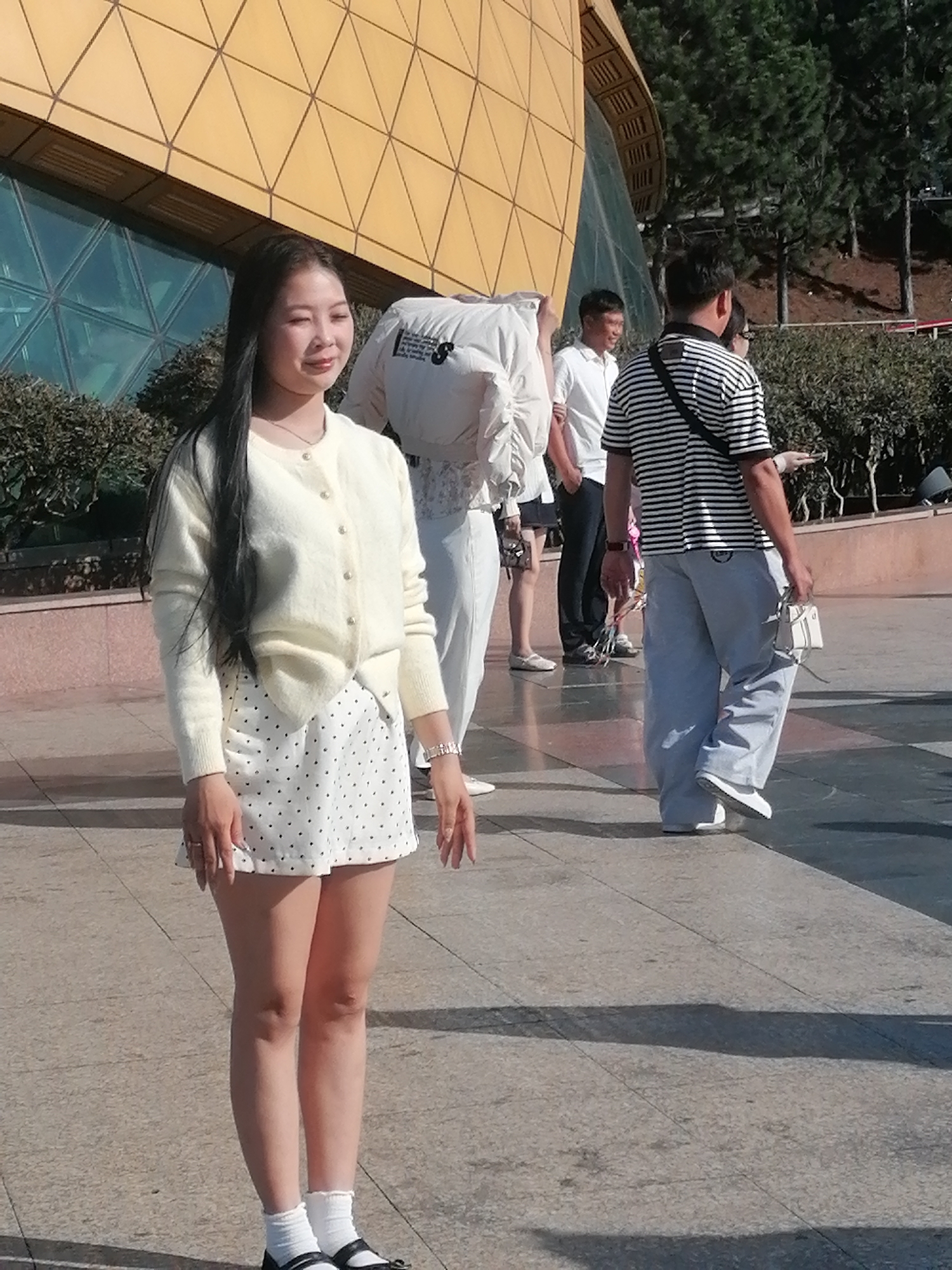In Da Lat, Vietnam, a young East Asian woman with long black hair stands in the foreground, wearing a cream-colored cardigan over a white polka-dot skirt. She has a delicate necklace and several rings on her fingers, and her feet are adorned with white socks and black Mary Jane shoes. Behind her, other individuals are visible, including a man in a black and white striped shirt and light blue pants carrying a small white purse, and another man in a white polo shirt. The background features a modern building with a distinctive yellow, geometric facade and lush green trees. The scene appears to be an outdoor public space, possibly a plaza or walkway, under bright sunlight, casting distinct shadows on the paved ground.