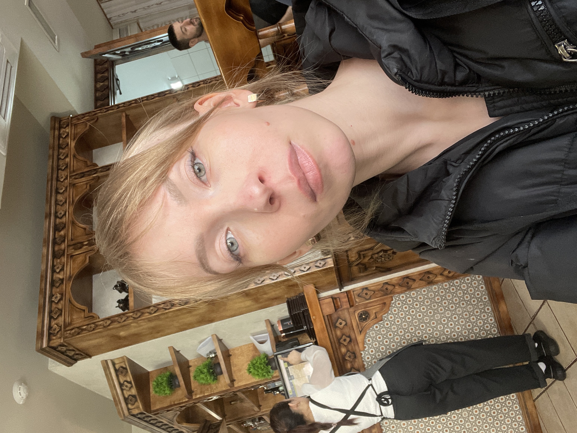The image depicts a blonde woman with fair skin and blue eyes in Perm, Russia. She is wearing a black jacket. In the background, there is a man reflected in a mirror. A waitress in a white shirt and black pants stands by a counter with plants and equipment. The interior features patterned tile flooring and decorative wooden shelves.