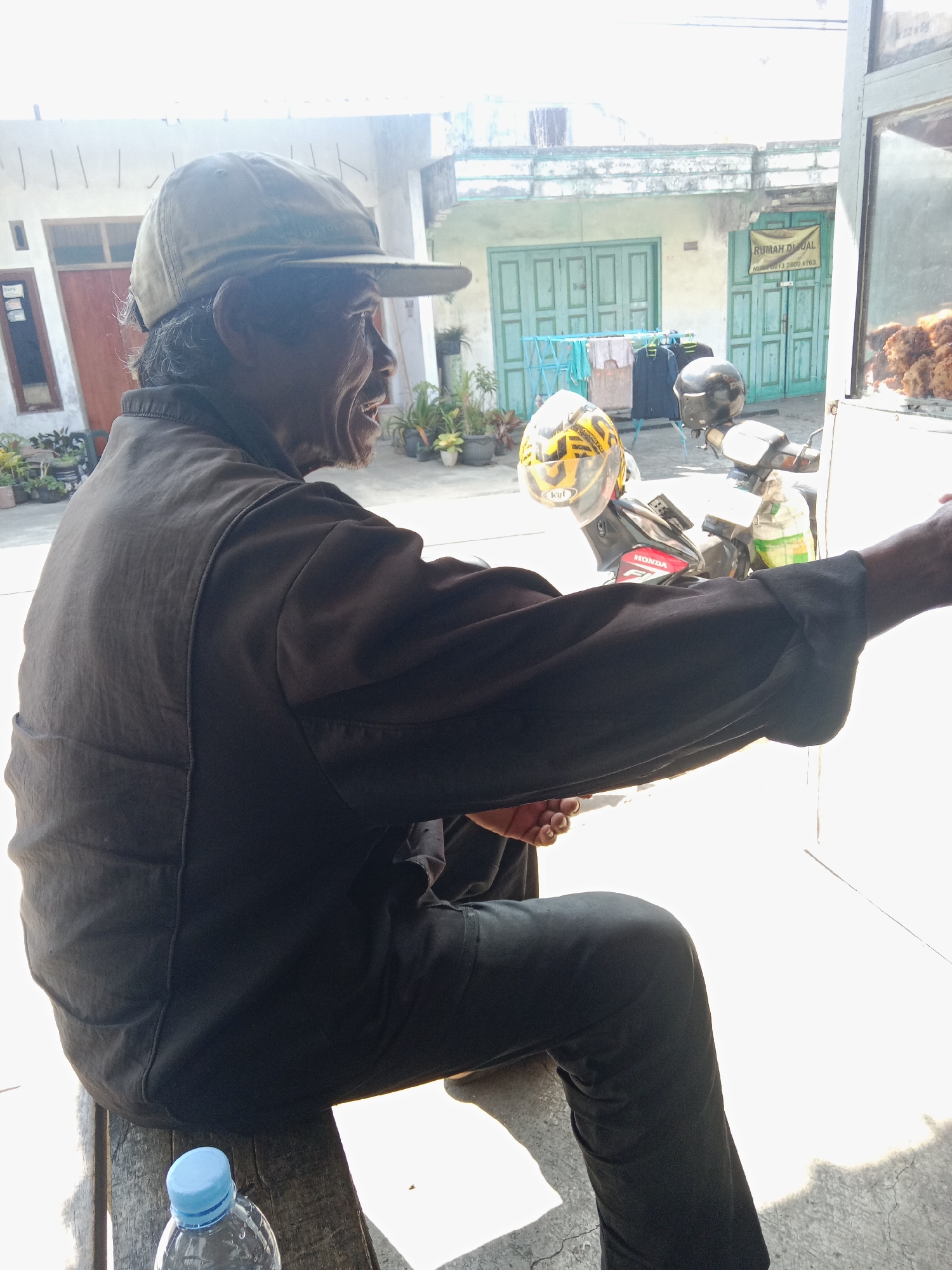 In Ringinanom, Indonesia, a man sits on a wooden bench under bright daylight. He is wearing a dark brown long-sleeved shirt, dark pants, and a light brown baseball cap. His body is angled to the right, and he appears to be speaking, with his mouth open. In front of him, to his left, is a plastic water bottle with a light blue cap. Behind him, a motorcycle with two helmets is parked. The background features a light-colored building with green doors and windows. A sign on the building reads "RUMAH DI JUAL" with a phone number. Laundry is hanging on a rack in the middle ground. The overall environment is outdoors with sunlight casting shadows.