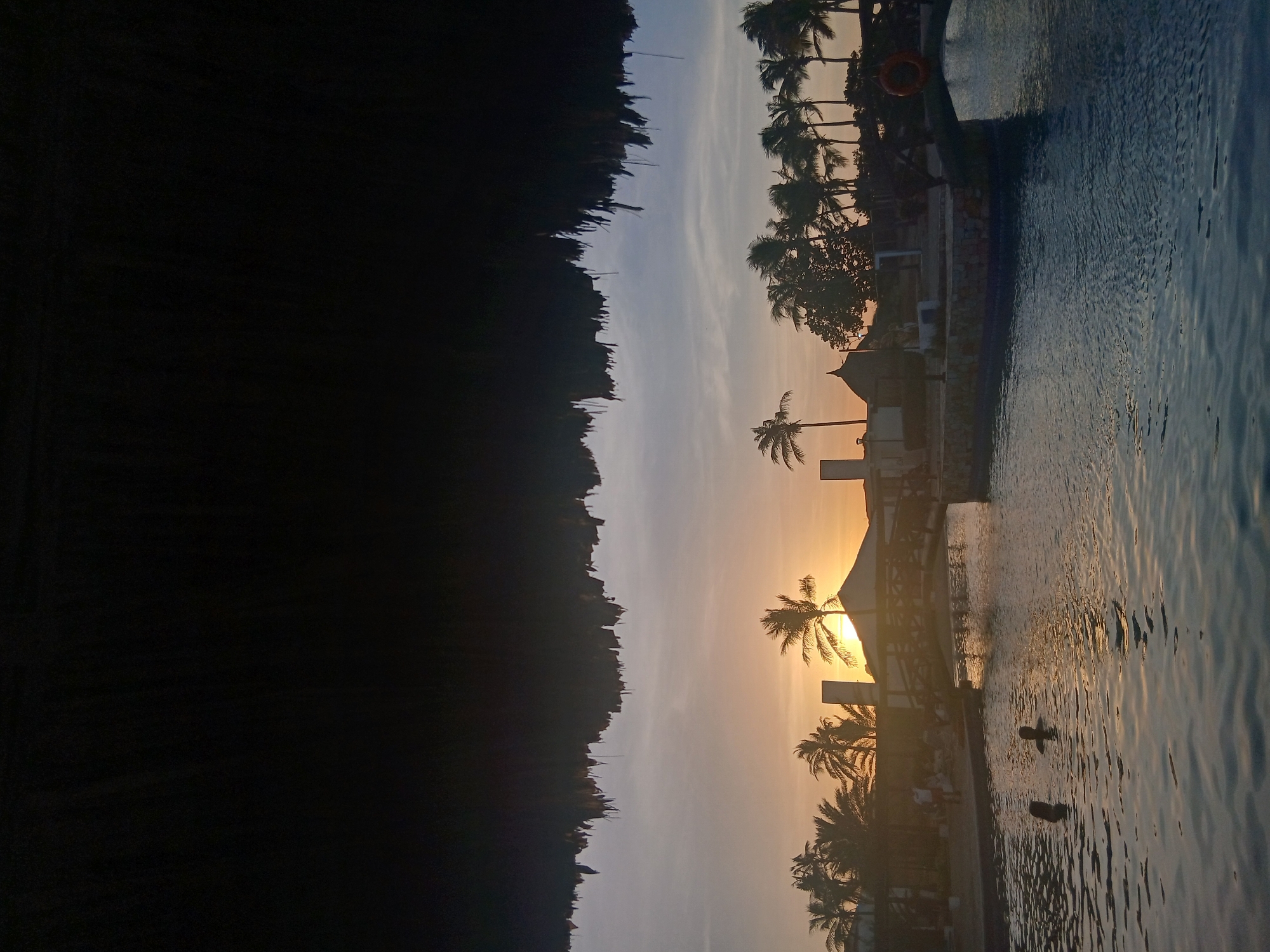 This image depicts a tropical resort scene at sunset in Altagracia, Venezuela, viewed from beneath a dark, fringed thatched roof that partially obscures the upper frame. A body of water, consistent with a large swimming pool or lagoon, occupies the lower half of the frame, showing ripples and reflecting the warm light of the setting sun. Two individuals are partially submerged in the water, their heads and upper shoulders silhouetted.
In the midground, an arched bridge, constructed with wooden railings and a stone base, spans a section of the water. A red circular life preserver is affixed to the right side of the bridge. Beyond the water and bridge, a land area features multiple structures, including buildings with distinctive peaked or thatched roofs and light-colored walls, interspersed among numerous palm trees. Several indistinct human figures are visible on this land area, appearing as silhouettes.
The background is dominated by the setting sun, positioned low on the horizon towards the center-left, emitting an intense orange-yellow glow that transitions to a pale blue-grey sky. Dense clusters of palm trees are silhouetted across the entire horizon, defining the tropical environment.