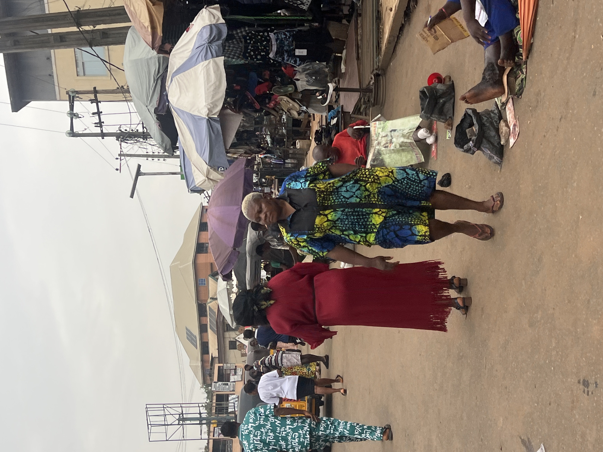 The image depicts a bustling market scene in Nkpor, Nigeria. Two women are in the foreground, one wearing a colorful blue and yellow patterned dress, and the other in a red dress. Behind them, a crowd of people move through the market area. Vendors sell goods under umbrellas and makeshift stalls. Clothes hang on racks. There are buildings and utility poles visible in the background, indicating an urban environment. The ground appears to be a dirt or unpaved surface. A person sits on the ground selling various items.