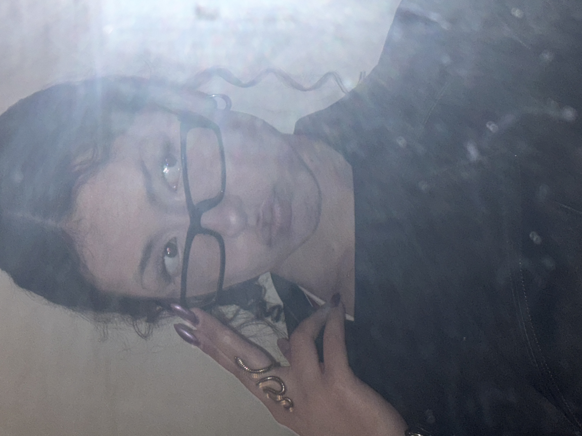 A woman with dark hair and glasses is positioned in front of a dimly lit background. She is wearing a dark-colored garment and has a snake-shaped ring on her finger. The image has a hazy, possibly fog-like quality, which obscures fine details. The location is not identifiable from the image.