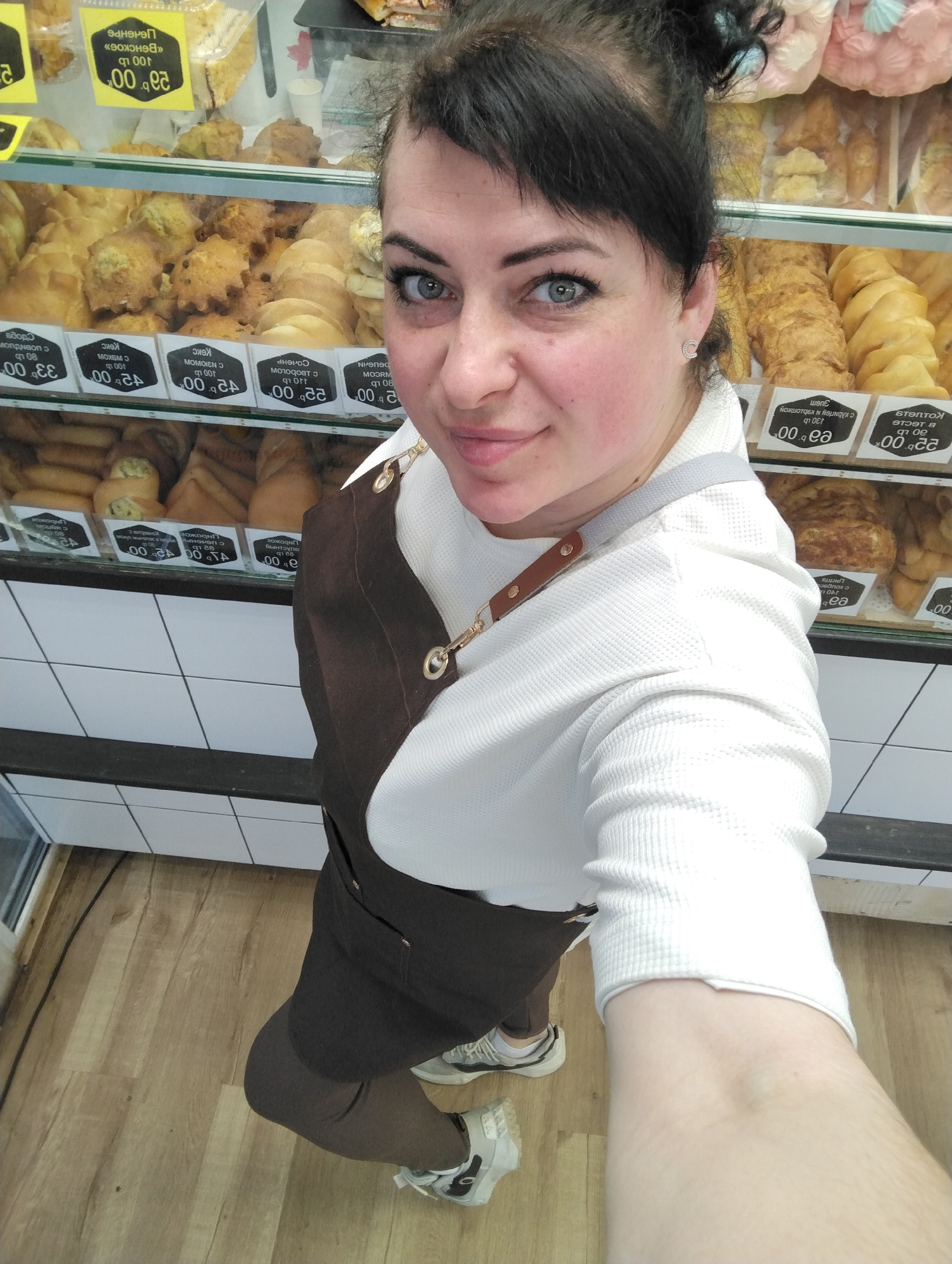 The image shows a woman in a brown apron and white long-sleeved shirt taking a selfie in front of a bakery display case. The case contains pastries and baked goods with price tags in Russian rubles. The location is the city of Votkinsk, Russia, as indicated by the language and currency on the price tags. The floor is wood-look vinyl. The wall beneath the bakery case is white tile. The woman is smiling and looking at the camera.
