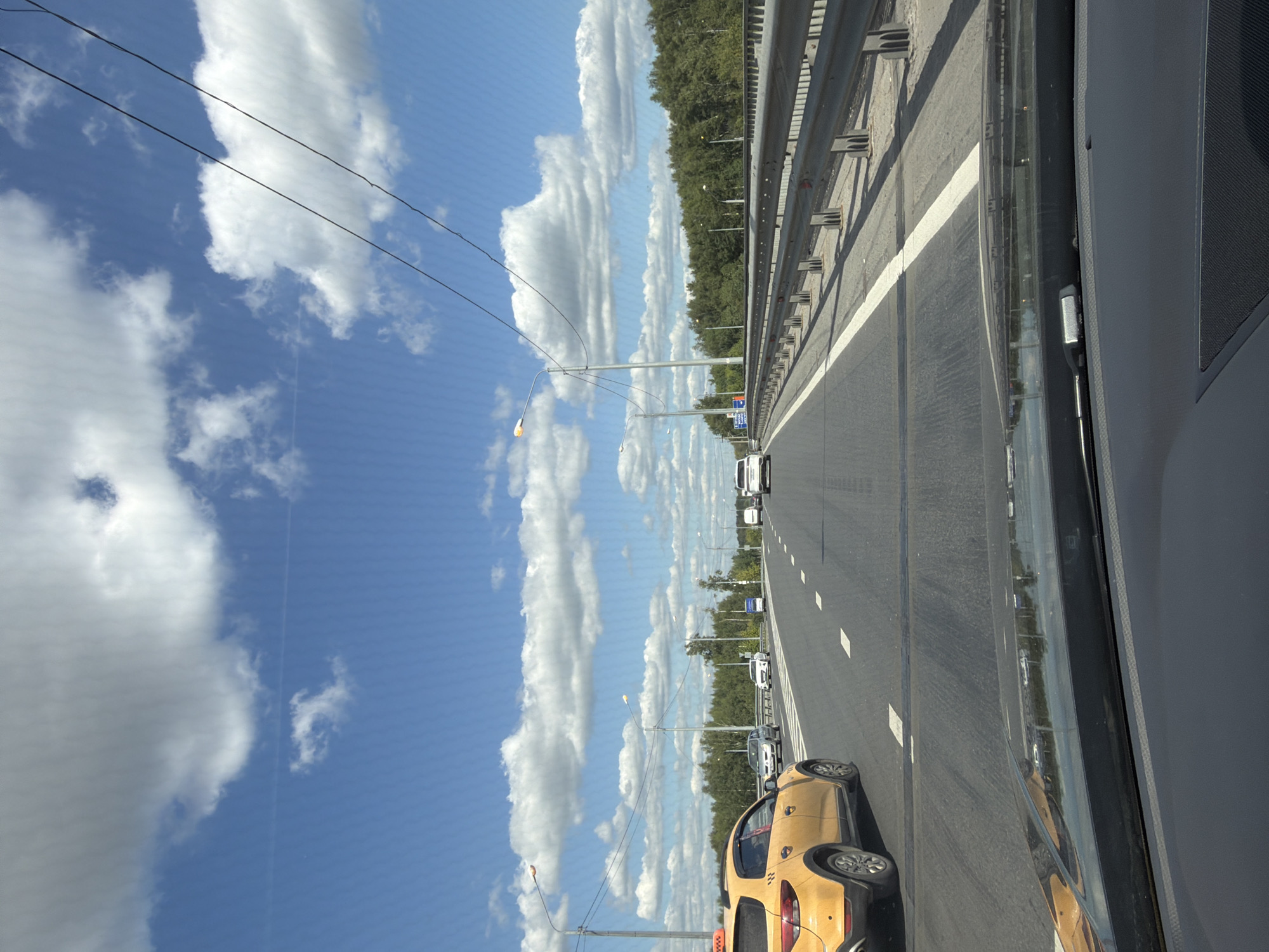 The image captures a view from the driver's seat of a vehicle on a highway, likely in Noginsk, Russia. The sky is bright blue with fluffy white clouds, suggesting fair weather. Power lines stretch across the sky. The highway has multiple lanes, with white lane markings. On the left side of the highway, a yellow taxi is visible in the foreground. Several other cars are driving on the highway in both directions. A metal guardrail lines the right side of the road, separating it from a densely wooded area. Streetlights are positioned along the highway. The dashboard of the vehicle in the foreground is visible at the bottom of the image.