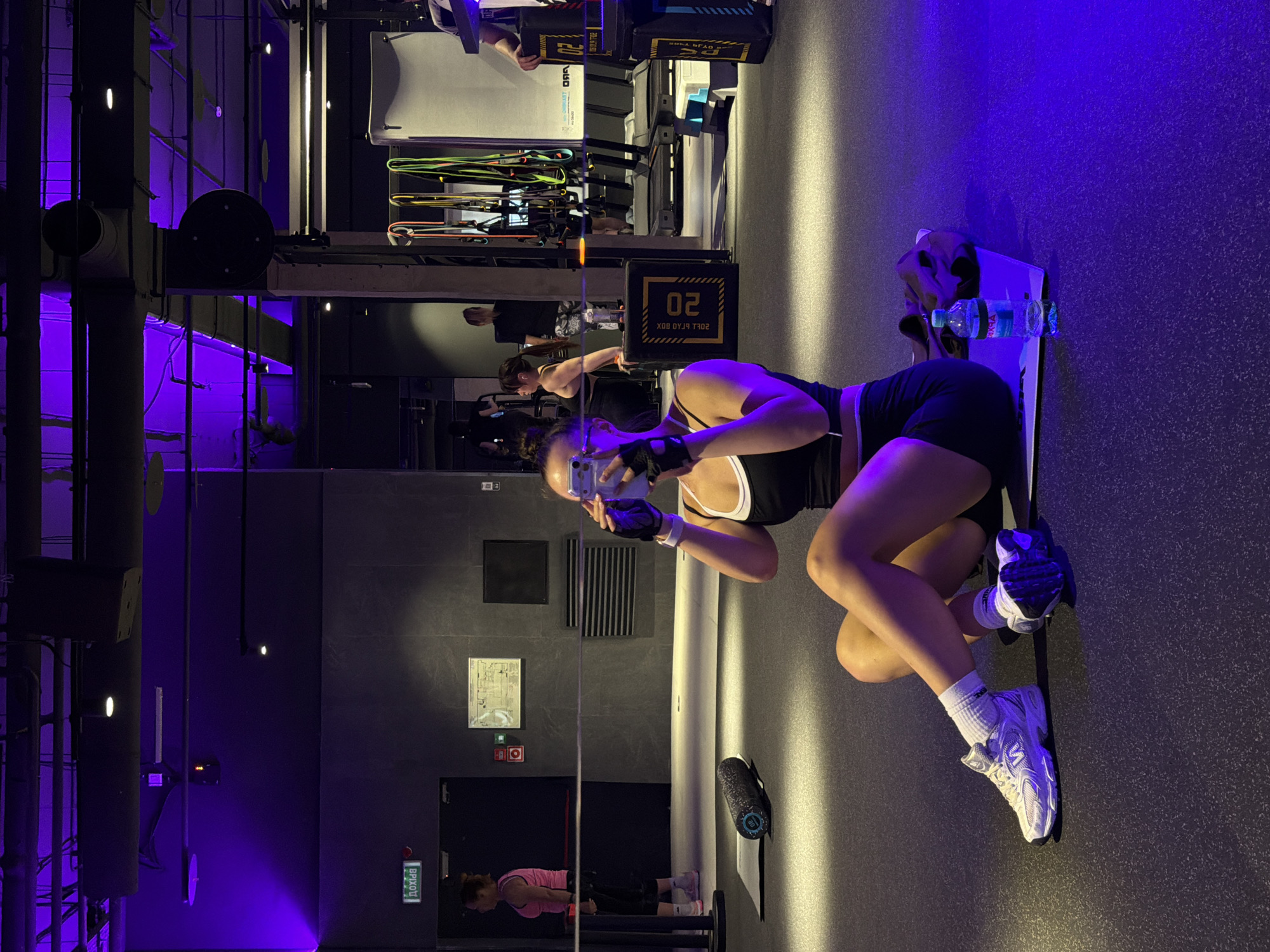 A woman in a black and white workout outfit is taking a selfie in a gym. She is wearing black athletic gloves and white athletic shoes. A water bottle is next to her on a bench. In the background, a woman is working out. The sign on the wall reads "Вход" and the emergency notification is in Russian; this indicates the scene is in Russia, likely Podolsk. The gym has blue and purple lighting.