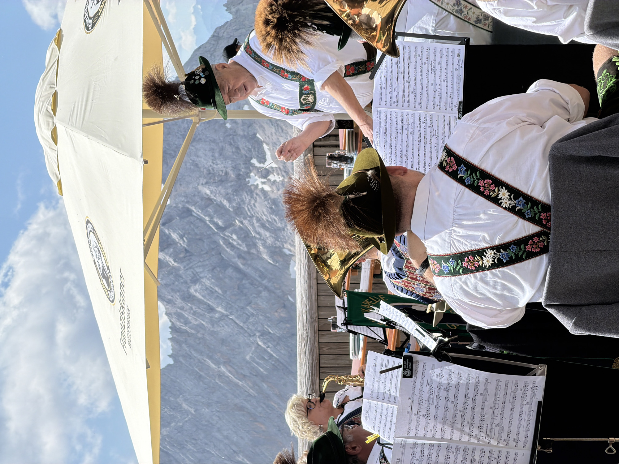 The image depicts an outdoor band performance under a large white umbrella advertising "Franziskaner Weißbier." In the foreground, several band members are visible, dressed in traditional Bavarian attire, including white shirts with embroidered suspenders and green hats adorned with feathers. One man, in the center, appears to be conducting the band with a baton. A woman is playing the saxophone to his left. Sheet music stands are set up in front of the musicians. In the background, a rugged mountain range is visible under a partly cloudy sky, suggesting a mountainous region. The overall scene evokes a festive, outdoor performance setting.