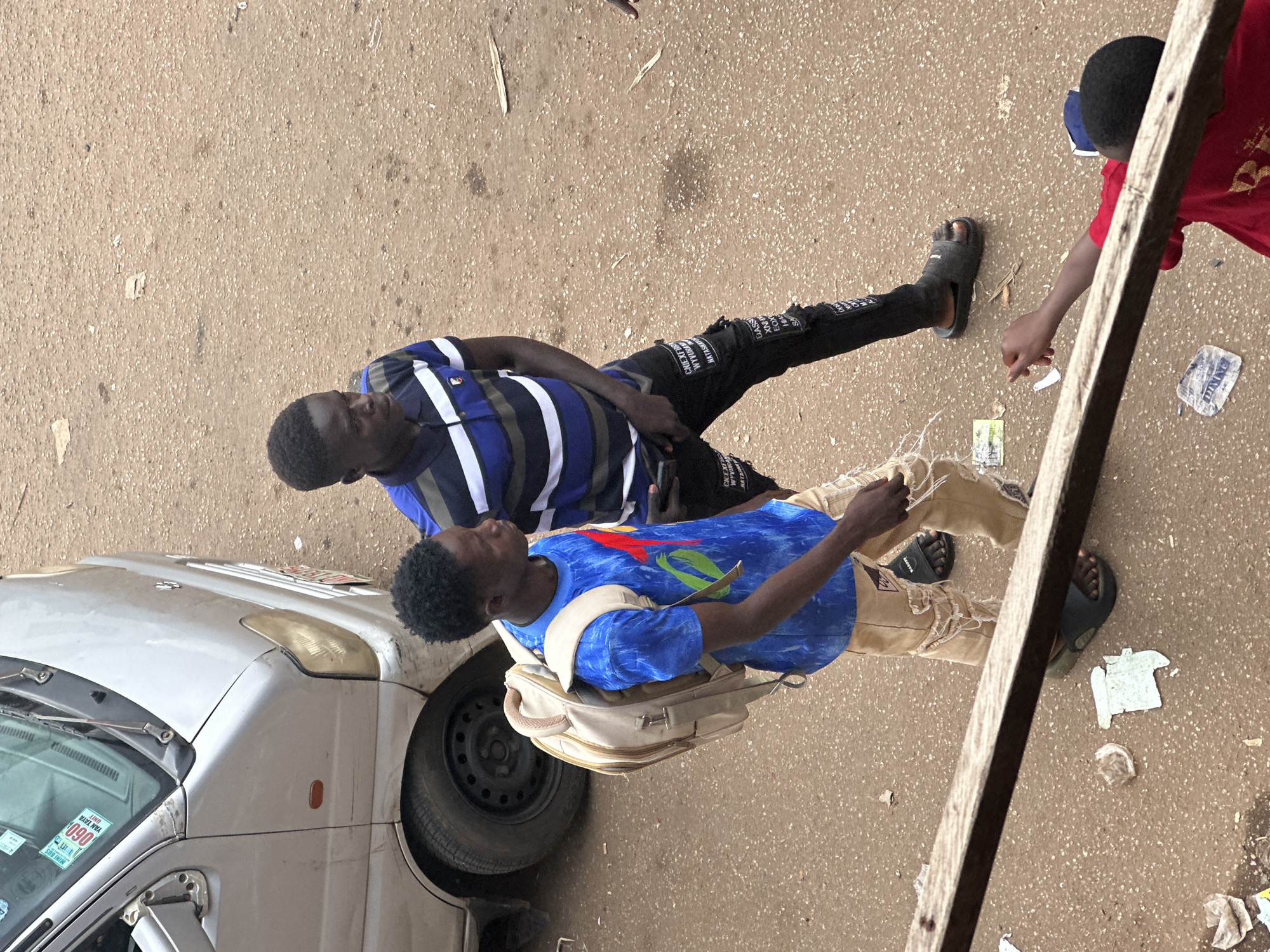 Two young men are walking in a street in Bukuru, Nigeria. The man on the left, wearing a blue t-shirt with a colorful graphic design, has a beige backpack on. He is also wearing distressed tan pants and black sandals. The man on the right is wearing a blue and white striped polo shirt and black pants with white text. They appear to be walking away from the camera. A silver van is parked to the left of the frame, and the ground is a dirt and gravel surface with scattered debris. A portion of a wooden plank is in the foreground. Another person in a red shirt is visible in the bottom right corner, reaching out.