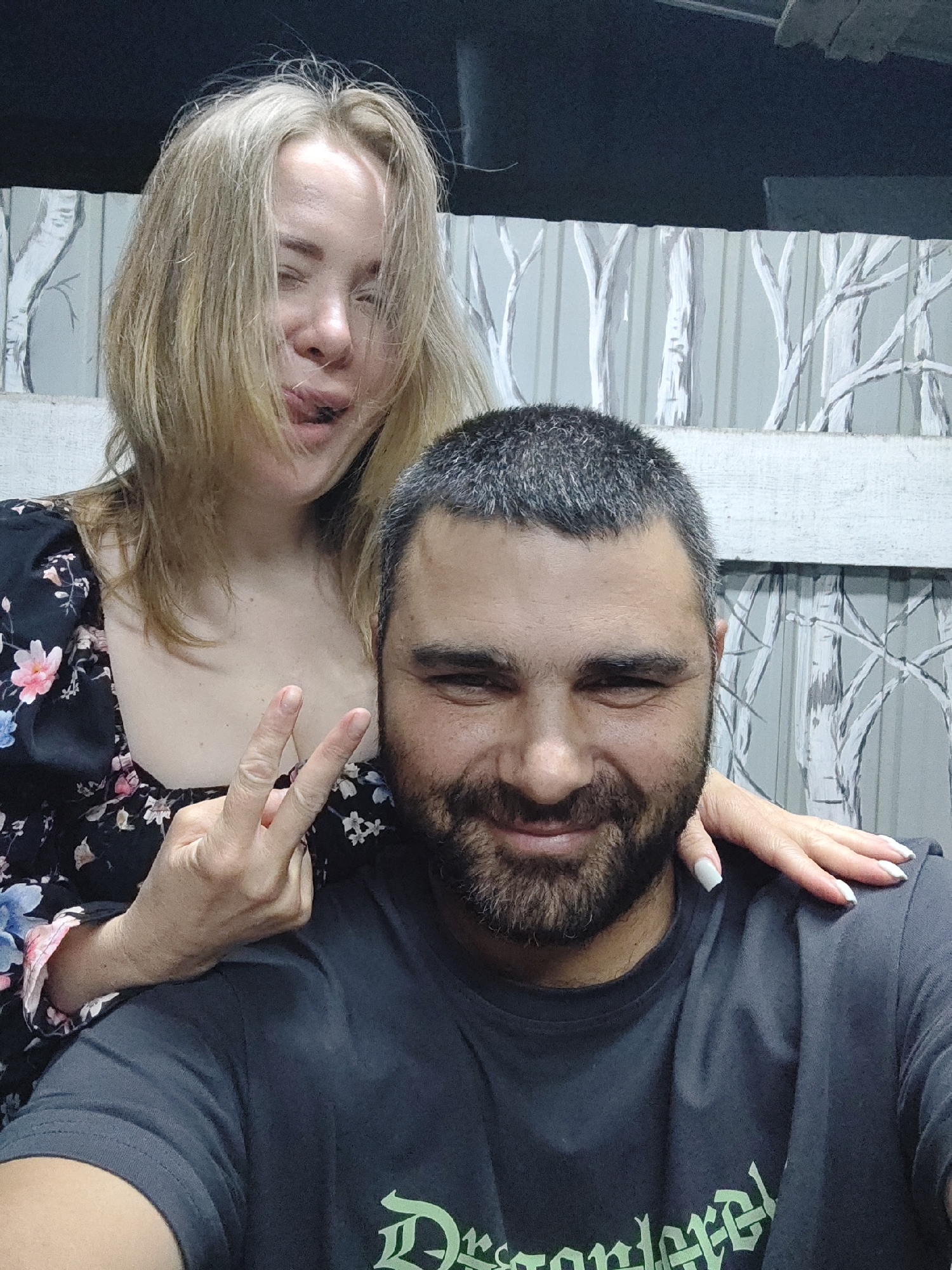 A close-up selfie shows a Caucasian man and woman. The man, in the foreground, has dark, short-cropped hair and a beard, and is looking forward with a slight smile, his right eye squinting. He wears a dark gray t-shirt. A woman with blonde hair is positioned behind and to the left of the man. Her face is partially obscured as she sticks her tongue out and squints her left eye. She is wearing a dark dress with a floral pattern. Her right hand is raised, fingers forming a peace sign, and is positioned in front of the man's chest. Her left hand rests on the man's right shoulder. The background features a wall with a repeating pattern of stylized, bare tree branches. The overall scene appears to be indoors, possibly in a residential setting, but the location in Shimanovsk, Russia cannot be determined from the image alone.