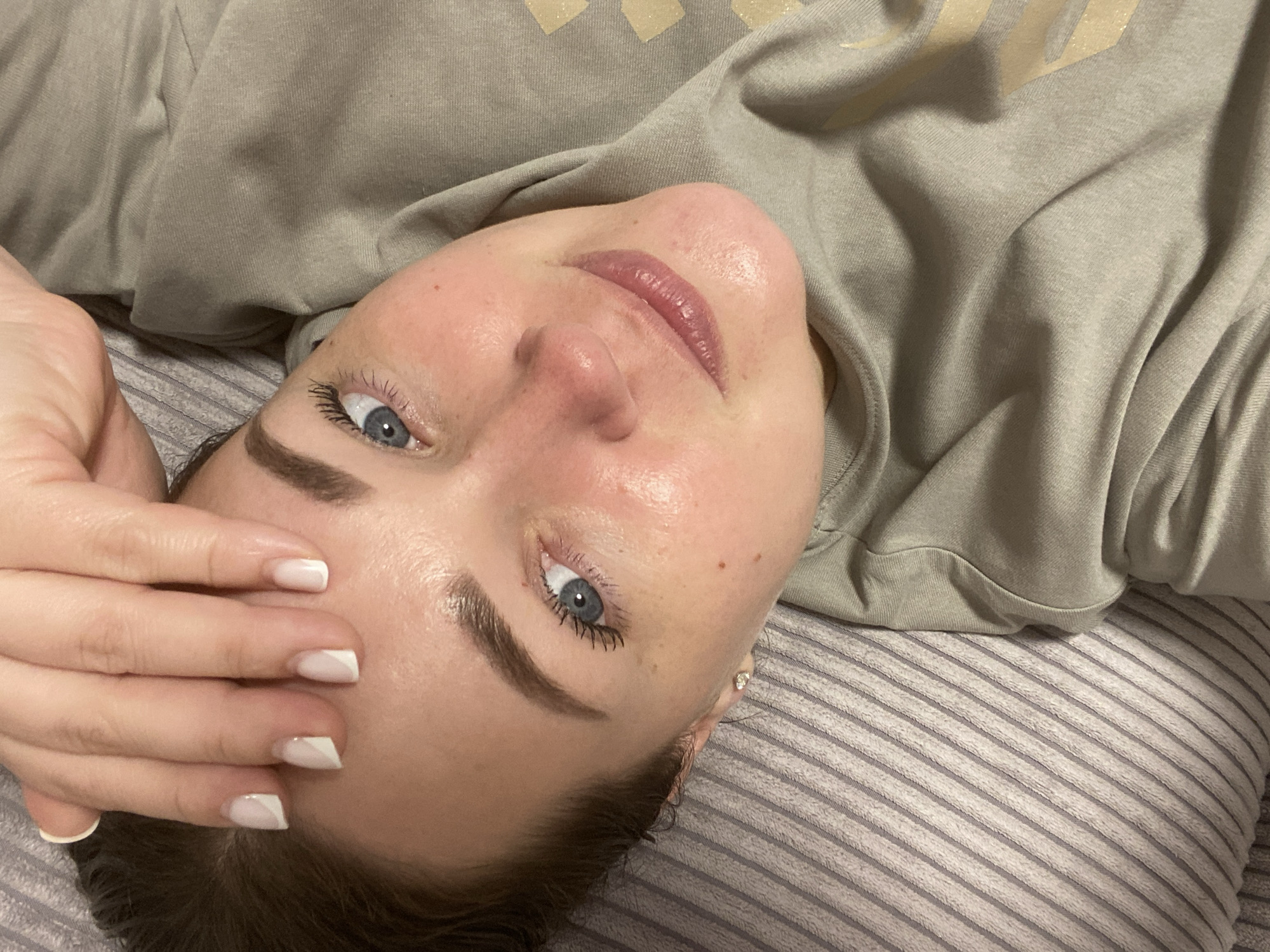 The image captures a close-up, high-angle portrait of a person, likely female, lying down indoors. The subject has clear blue eyes, defined dark eyebrows, and a relaxed expression with a hint of a smile. Her skin appears healthy and slightly dewy, and she wears light-colored lipstick. A small, shiny stud earring is visible in her left ear.
She is wearing a light olive-green or beige long-sleeved top or sweatshirt, which has a subtle, lighter-colored design (possibly text or a logo) on the chest area, though it is not legible. Her left hand is resting on her forehead, displaying manicured nails with a white French tip design, some featuring a diagonal style.
The person is lying on a surface covered by a soft, striped fabric, likely a pillow or blanket, which features alternating light and darker grey stripes. The setting appears to be an indoor environment, possibly a bedroom or living room, with soft, ambient lighting. The overall mood is calm and intimate. There are no discernible text elements beyond the unreadable logo on the shirt. No specific geographical location can be identified from the image.