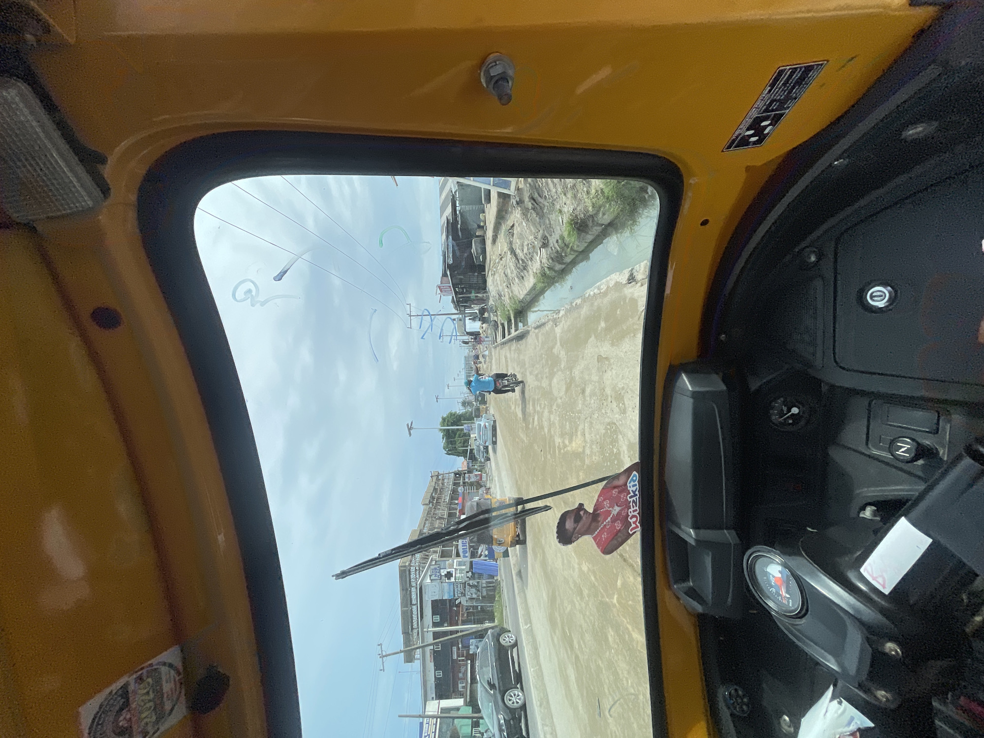 A view from inside a yellow three-wheeled vehicle, possibly an auto-rickshaw, in Ado, Nigeria. Through the front windshield, a street scene is visible. The street appears to be paved, and there's a waterway running along the side of the road. There are buildings on both sides of the street, with some under construction. Various vehicles, including cars and a motorcycle, are on the street. A wiper blade is visible on the windshield, and a small figure with the name "Wizkid" attached to it is placed on the wiper blade. The interior of the auto-rickshaw shows a dashboard with gauges and controls. The sky is overcast.