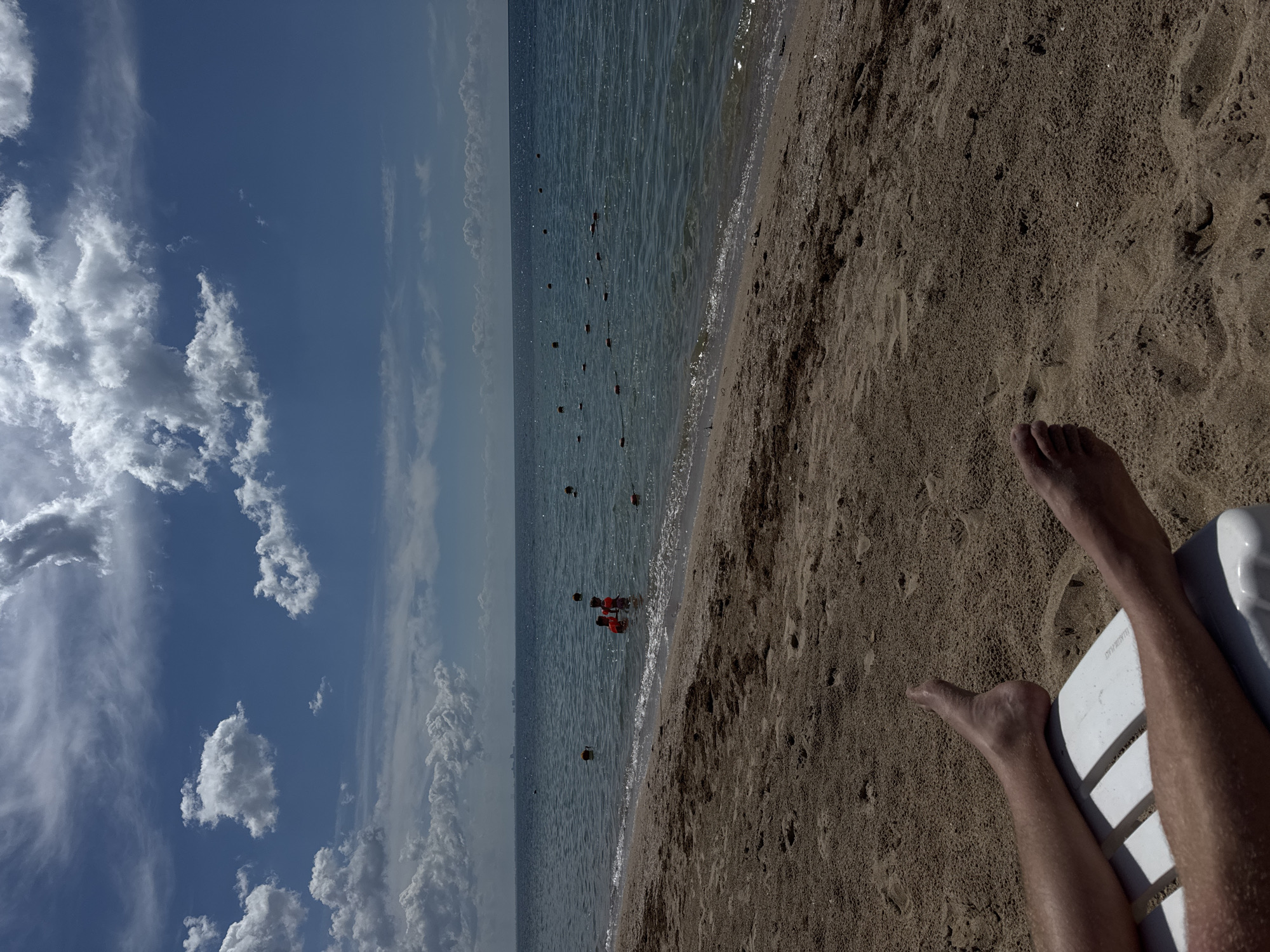 The image depicts a coastal scene, possibly in the city of Yevpatoriya, Ukraine. The view is predominantly horizontal, featuring a light-blue sky with scattered white clouds occupying the left portion. The sky transitions into a seascape with gentle waves. In the water, there are several floating buoys and a group of people wearing what appears to be red life vests, close to the shoreline. The right side of the frame is composed of a sandy beach. A pair of human legs and feet, likely belonging to someone reclined on a white plastic lounge chair, occupy the lower right corner. The overall atmosphere suggests a sunny, relaxed beach setting.