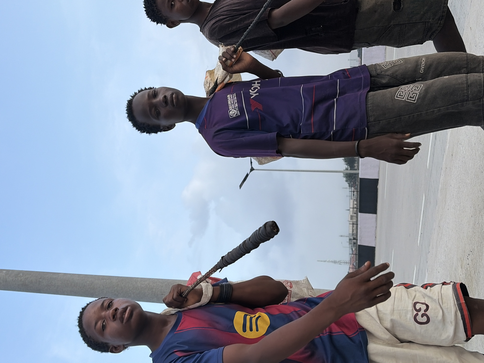 Three young boys, appearing to be in their early to mid-teens, are walking on a paved road in Lekki, Nigeria. The boy on the left wears a blue and red striped t-shirt with a yellow Spotify logo on the chest, and light-colored shorts with a Gucci logo. He is carrying a bundle wrapped in white fabric over his left shoulder, from which a dark, thick rope extends, held in his right hand. The middle boy wears a purple t-shirt with white and red stripes and a logo that reads "GEORGIAN GLOBAL GLOBAL GROUP" and "YOH". He is also carrying a bundle over his left shoulder with a rope in his right hand. He wears dark pants with a white geometric pattern and "GIVENCHY" text. The boy on the right is partially visible, wearing a dark t-shirt and dark shorts, and appears to be carrying something similar. They are walking on a road with clear lane markings and a black and white striped barrier along the side. In the background, there are some buildings and a tall structure resembling a communication tower, suggesting an urban or semi-urban environment. The sky is overcast with some clouds.