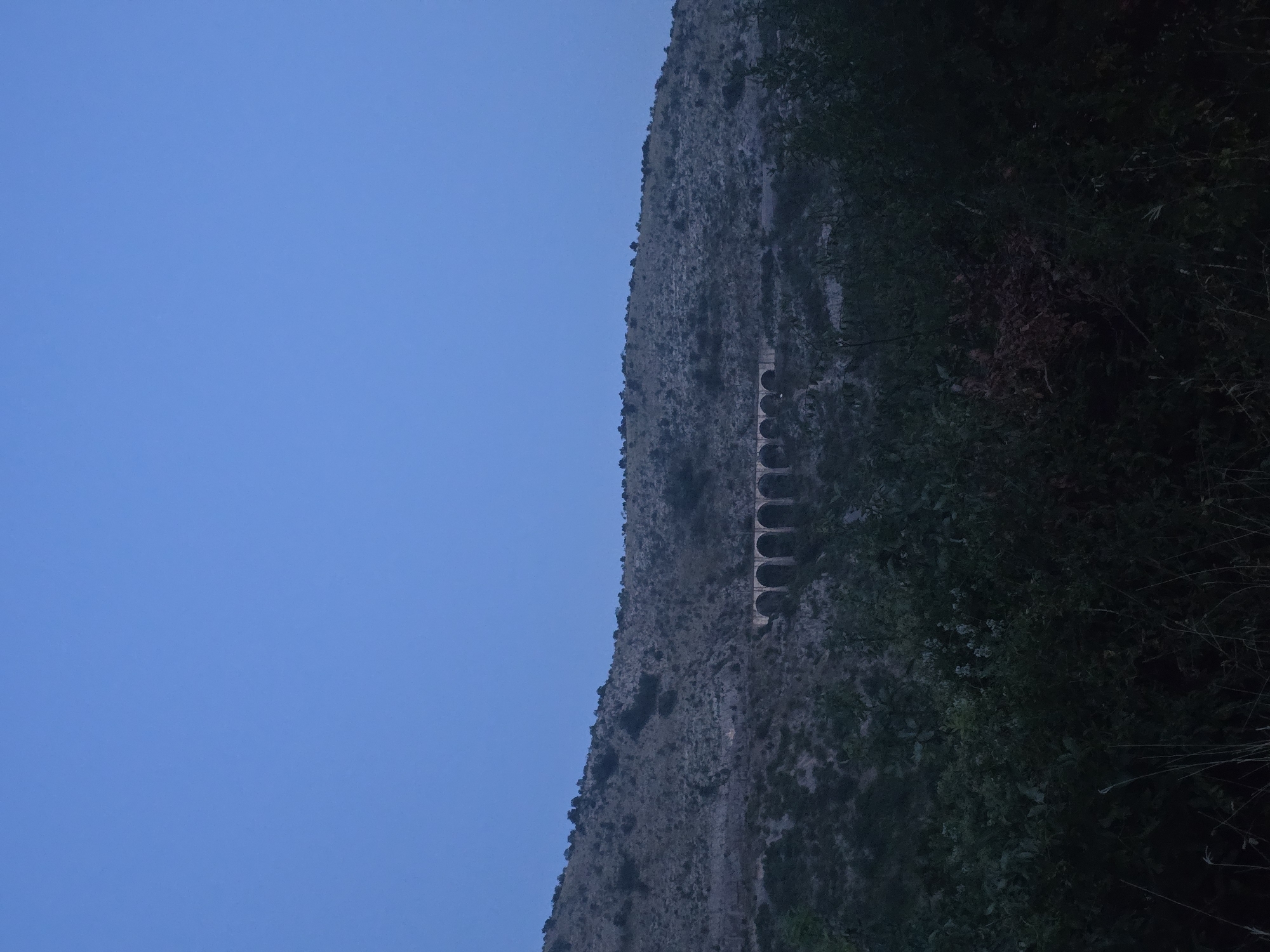 The image shows a wide shot of a hilly landscape at dusk. In the foreground, dense, dark green foliage obscures the lower portion of the scene. In the middle ground, a mountainous incline covered in sparse vegetation rises towards the sky. A stone aqueduct, characterized by a series of arches, traverses the hillside horizontally, appearing weathered and aged. The aqueduct is illuminated by the fading light. The sky above is a deep, uniform blue, indicating the transition from day to night. No people or specific structures beyond the aqueduct are visible. The location is identified as Klopot, Montenegro, suggesting a natural or semi-natural environment, likely rural or mountainous.