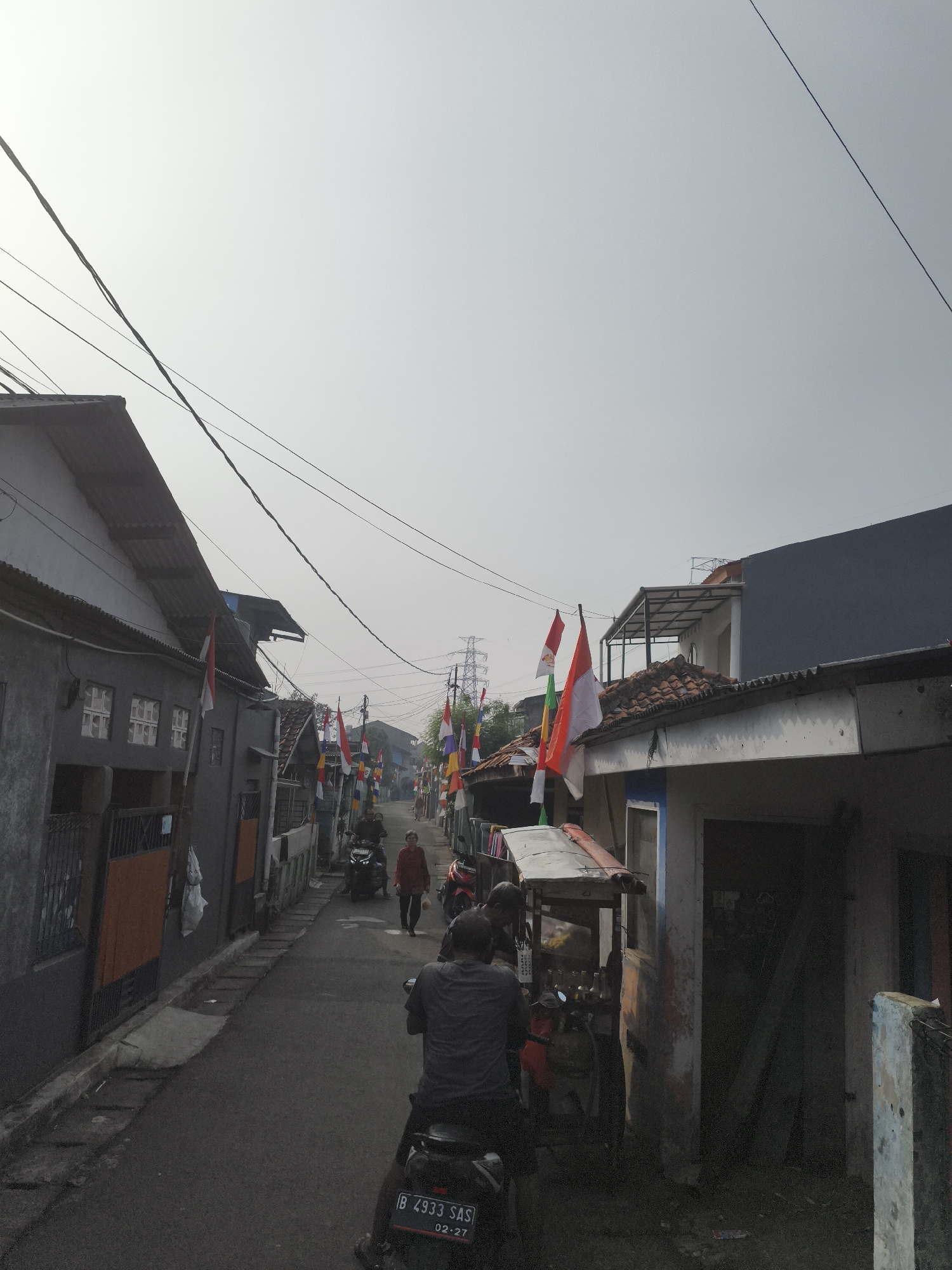This is a wide, slightly low-angle, eye-level shot of a narrow street in a residential area of Depok, Indonesia, possibly during a national holiday given the abundance of Indonesian flags decorating the houses and poles along the street. The sky is overcast and bright, with power lines crisscrossing overhead.
On the left side of the street, there are two-story houses with gray stucco walls and corrugated metal roofs. Some windows are visible, and the entrance to one of the houses features a dark metal gate.
The street itself is paved and appears to be mostly empty, with a slight incline. In the middle ground, a woman in a red jacket is walking away from the camera, and a man on a motorcycle is riding towards her. Further down the street, more Indonesian flags are visible, as well as a large electricity pylon in the distance.
On the right side of the street, there are more houses, some with tiled roofs. In the foreground, a man is sitting on a motorcycle with a license plate reading "B 4933 SAS". Next to him is a small street food cart with bottles of drinks displayed. The cart is under a makeshift awning.
The overall impression is of a community preparing for or celebrating a local event, with a sense of daily life unfolding in a densely populated urban environment.