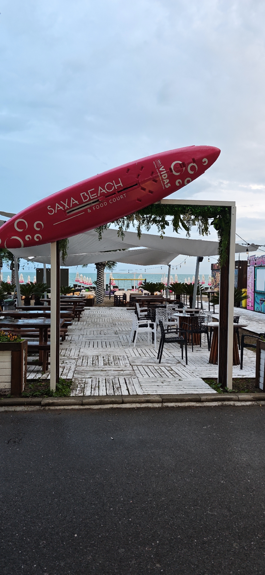 The image shows the entrance to "Saxa Beach & Food Court" in Burgas, Bulgaria, identifiable by the signage on a large pink, airplane-shaped structure atop the entrance. The entrance is framed by a square arbor-like structure with green plants. Inside the entrance, a white wooden deck extends into the dining area with tables and chairs. In the background, beach umbrellas and the sea are visible. The foreground consists of a paved road. The sky is overcast. There are no people visible in the image.