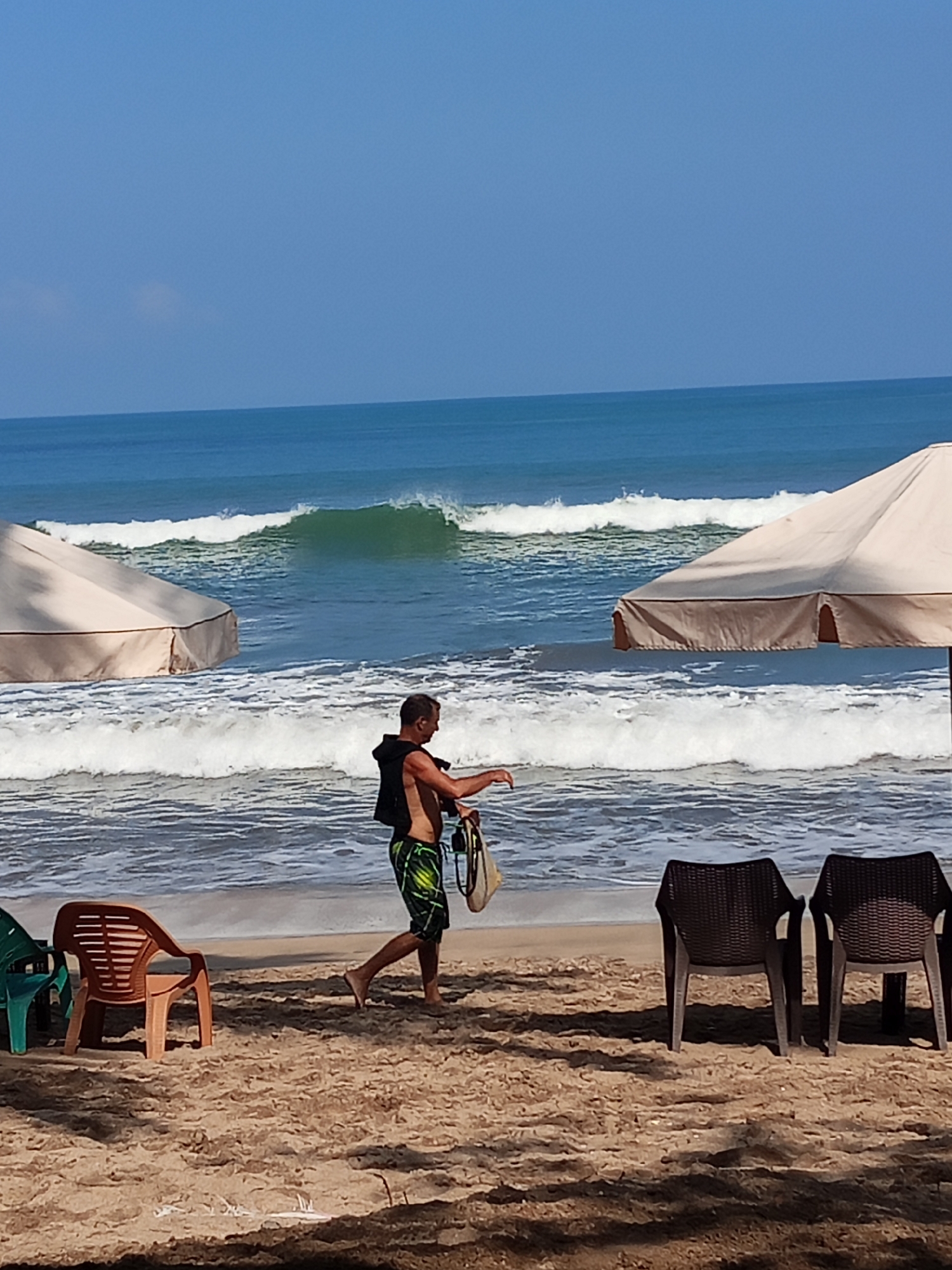 The image depicts a beach scene in Kuta, Indonesia, under a clear blue sky. In the mid-ground, a man wearing a black vest over a patterned swimsuit walks on the wet sand near the shoreline. He is carrying a round object, possibly a disc or a small board, in his right hand. In the background, the ocean features breaking waves with a prominent green crest. Two beach umbrellas are visible, one on the left and another on the right, providing shade. Several plastic chairs, including a green one and an orange one, are arranged on the sand in the foreground, along with two dark-colored chairs on the right. The sand is light brown and appears dry in the foreground, with shadows cast from the umbrellas and chairs.