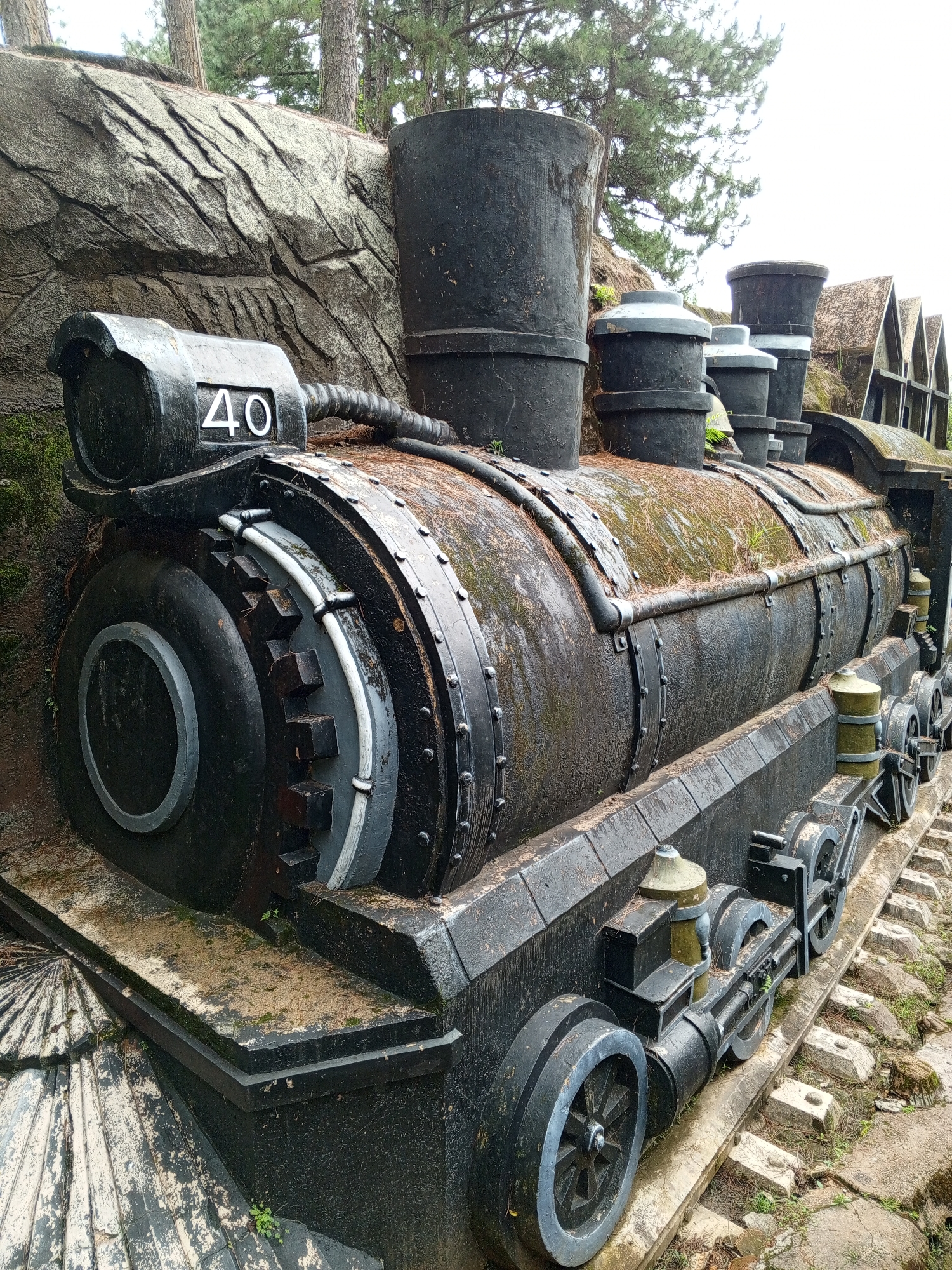 The image shows a weathered, black, stylized replica of a steam locomotive, made of concrete or a similar material. The replica is situated outdoors, with a textured rock face behind it and a few pine trees and a cloudy sky visible in the background. Several small, triangular-shaped buildings with brown roofs are visible on the right side of the image, behind the locomotive. The locomotive replica is the primary subject, detailed with wheels, pipes, and a prominent number "40" displayed on its front section. The surface of the locomotive is covered with moss and pine needles, indicating it has been exposed to the elements for some time. The replica appears to be a static display, possibly a tourist attraction. Based on the presence of pine trees and the style of architecture, this scene is likely located in Da Lat, Vietnam, known for its pine forests and French colonial-era architecture.