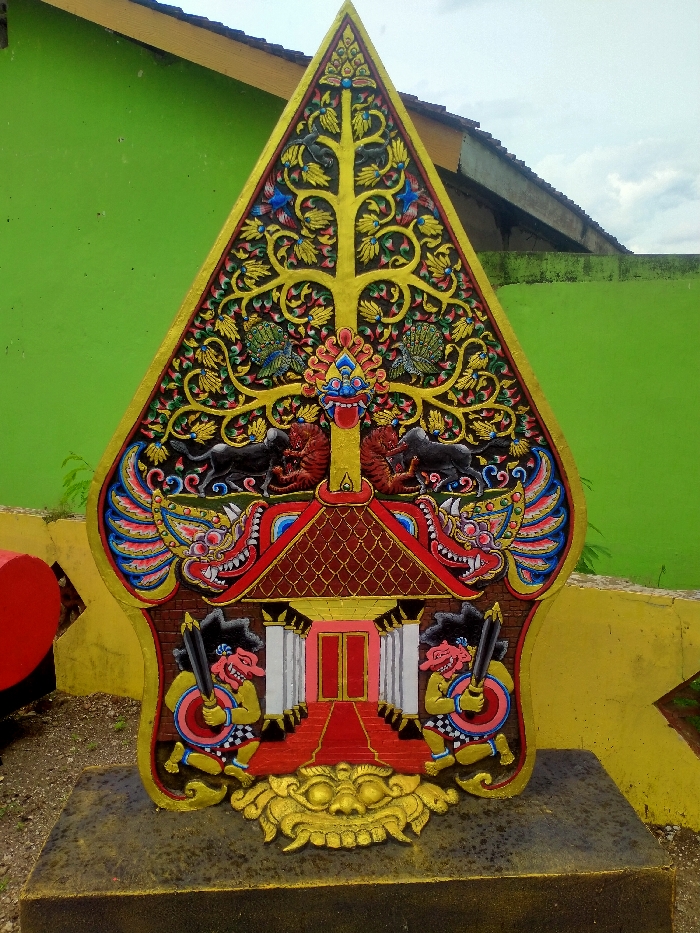 A colorful shadow puppet representation stands prominently outdoors. It features a golden tree of life adorned with blue, red, and gold details. Below, mythical creatures and black animals flank a red roofed structure with white columns. Two figures with clubs stand guard at the base, above a golden mask. The artwork is mounted on a gray base, set against a bright green wall.