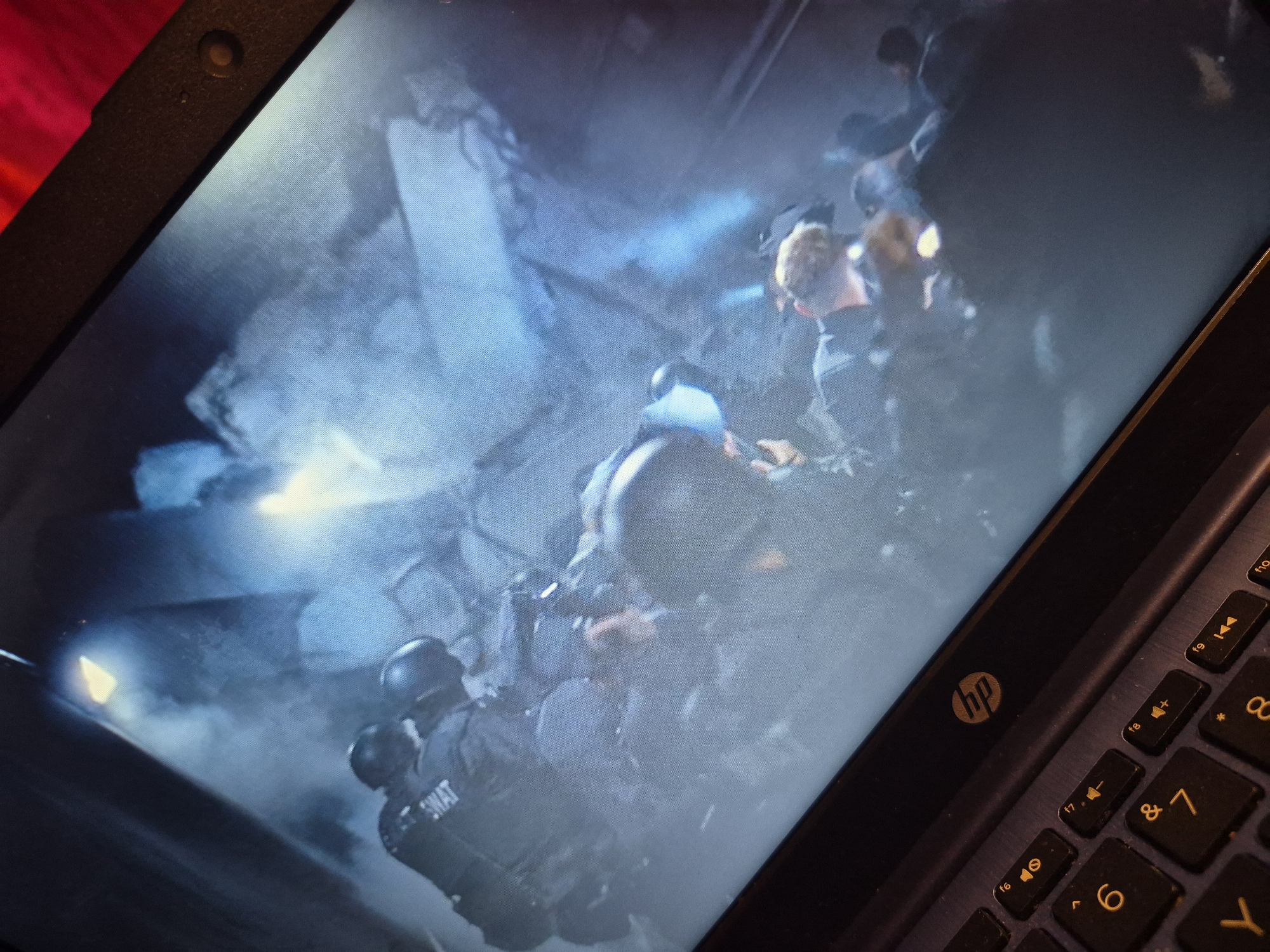 The image shows a laptop screen displaying a scene from a movie or show. The screen depicts several individuals in tactical gear, possibly SWAT officers, in a dimly lit, debris-filled environment with smoke or fog. One officer has "SWAT" visible on their uniform. The overall atmosphere is chaotic and suggests an ongoing operation or aftermath of an event. The laptop itself is an HP model, with visible keys including F keys, numbers, and letters. The surrounding area is out of focus, but appears to be an indoor setting. There is no discernible information within the image to identify a specific location in Sandwell, United Kingdom.