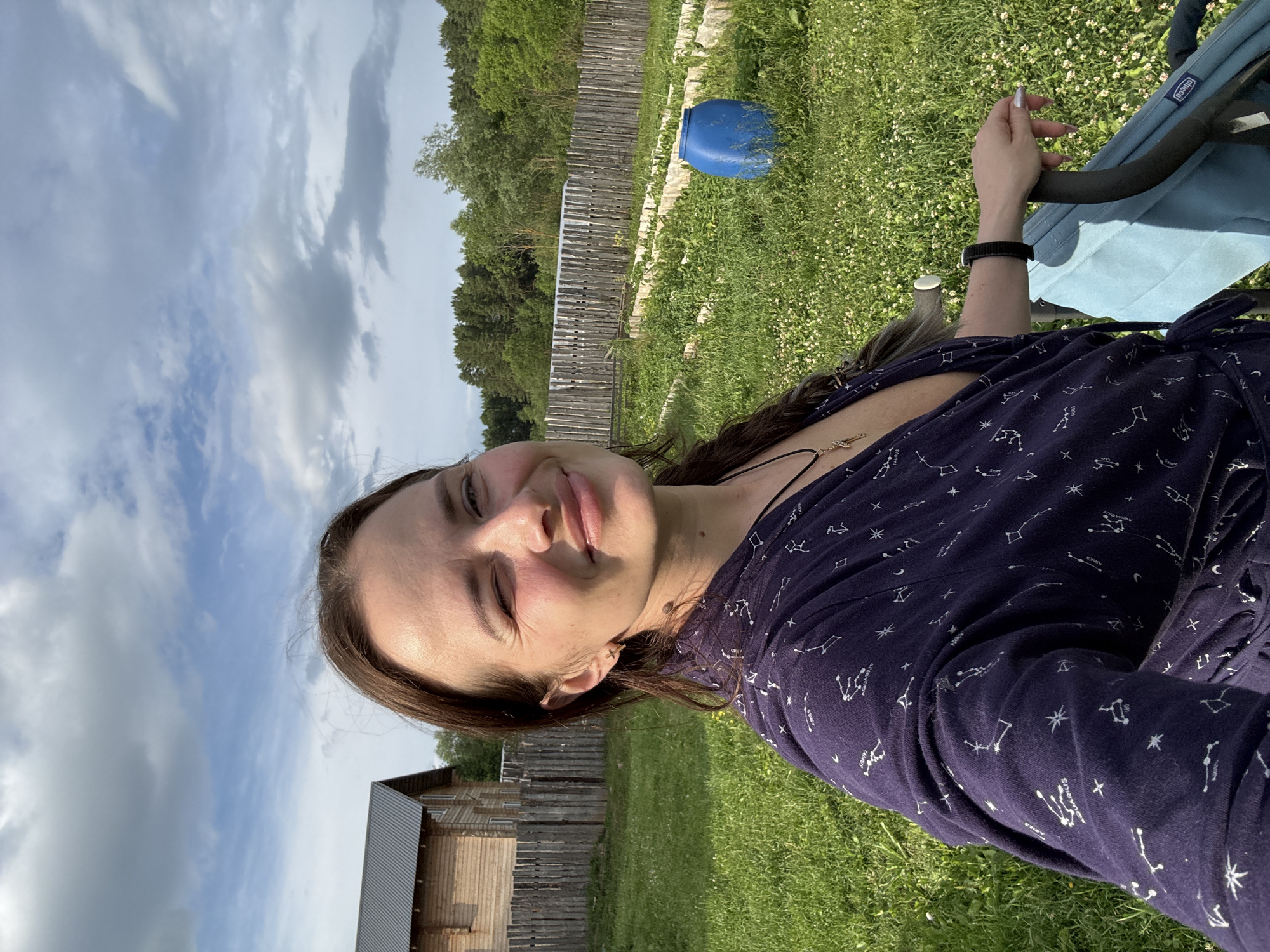 A woman is taking a selfie outdoors in what appears to be the rural village of Rusalevka, Russia. She is wearing a dark blue robe with white constellations printed on it. Her right eye is closed in a wink, and she is smiling. She has long, dark brown hair, and is wearing a gold cross necklace and a black smartwatch. Her left hand is resting on the handlebar of a light blue stroller. In the background, there is a wooden house with a dark roof, a wooden fence, a garden with raised beds, a blue barrel, and lush green grass and trees. The sky is partly cloudy with blue patches. The overall scene suggests a casual, possibly maternal, moment in a rural setting.