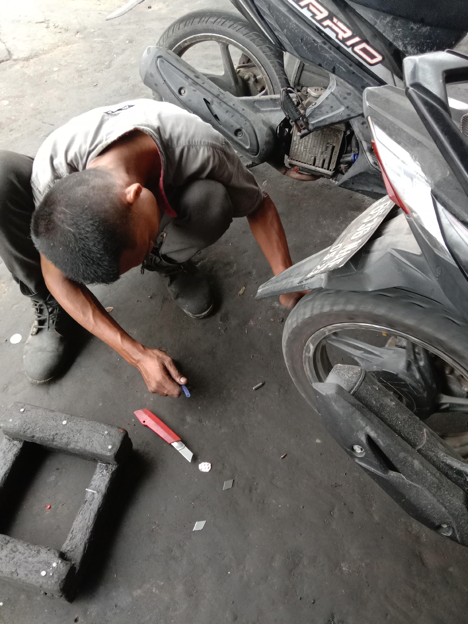 In a dimly lit garage in Lhokseumawe, Indonesia, a mechanic crouches near a black motorcycle. The motorcycle has the word "VARIO" visible on its side. The mechanic, dressed in a grey shirt and dark pants, is focused on a task near the rear tire. Scattered on the dirty concrete floor are tools, including a red box cutter, and small metal pieces. A makeshift frame-like object rests on the floor to the left. The scene depicts a typical motorcycle repair environment.