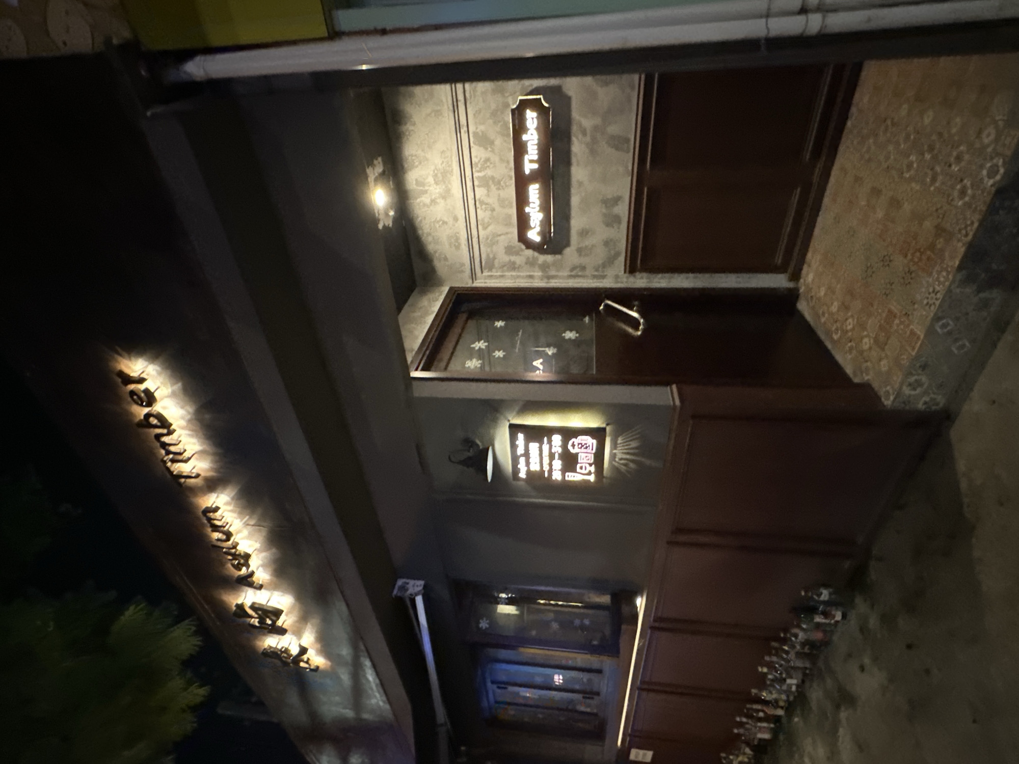 The image shows the exterior of a building at night, identified as "Asylum Timber" and located in the Chongqing High-tech Zone, China. The building features a dark facade with illuminated signage. The main sign above the entrance reads "Asylum Timber" in English, with additional text in Chinese characters that are not legible. A smaller, illuminated sign on the wall to the right of the entrance displays what appears to be opening hours in Chinese, along with icons of a glass and a beer mug. The entrance itself is a dark wooden door with a metal handle, and the surrounding wall has a textured, greyish finish with dark wooden paneling below. To the left of the entrance, a window with frosted snowflake-like designs is visible. The ground area in front of the building is partially visible, showing a tiled entrance step and a row of small bottles lined up along the dark wooden wall. The lighting suggests an evening or nighttime setting, with the illuminated signs being the primary light sources. There are no people visible in the scene. The overall impression is of a dimly lit, possibly commercial establishment, perhaps a bar or restaurant.