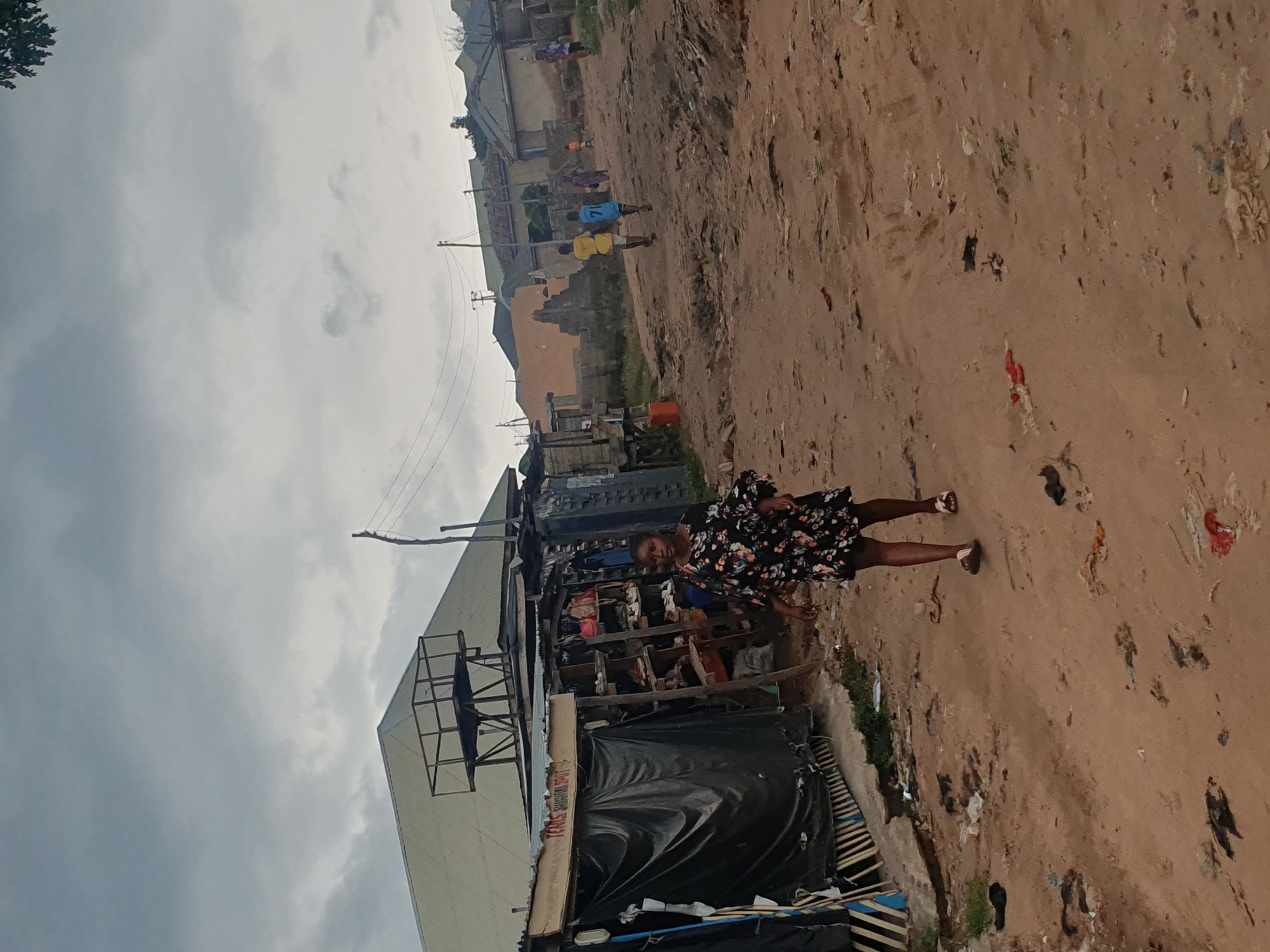 A young woman wearing a dark floral print dress walks on a dirt road in Lokoja, Nigeria. The road is unpaved and littered with debris. On the left, there are makeshift stalls with black tarpaulin coverings, displaying various goods on shelves. Behind the stalls, a large building with a sloped roof and an elevated metal platform is visible. In the background, more buildings and a few scattered figures, including children, are seen along the road. The sky is overcast with gray clouds.