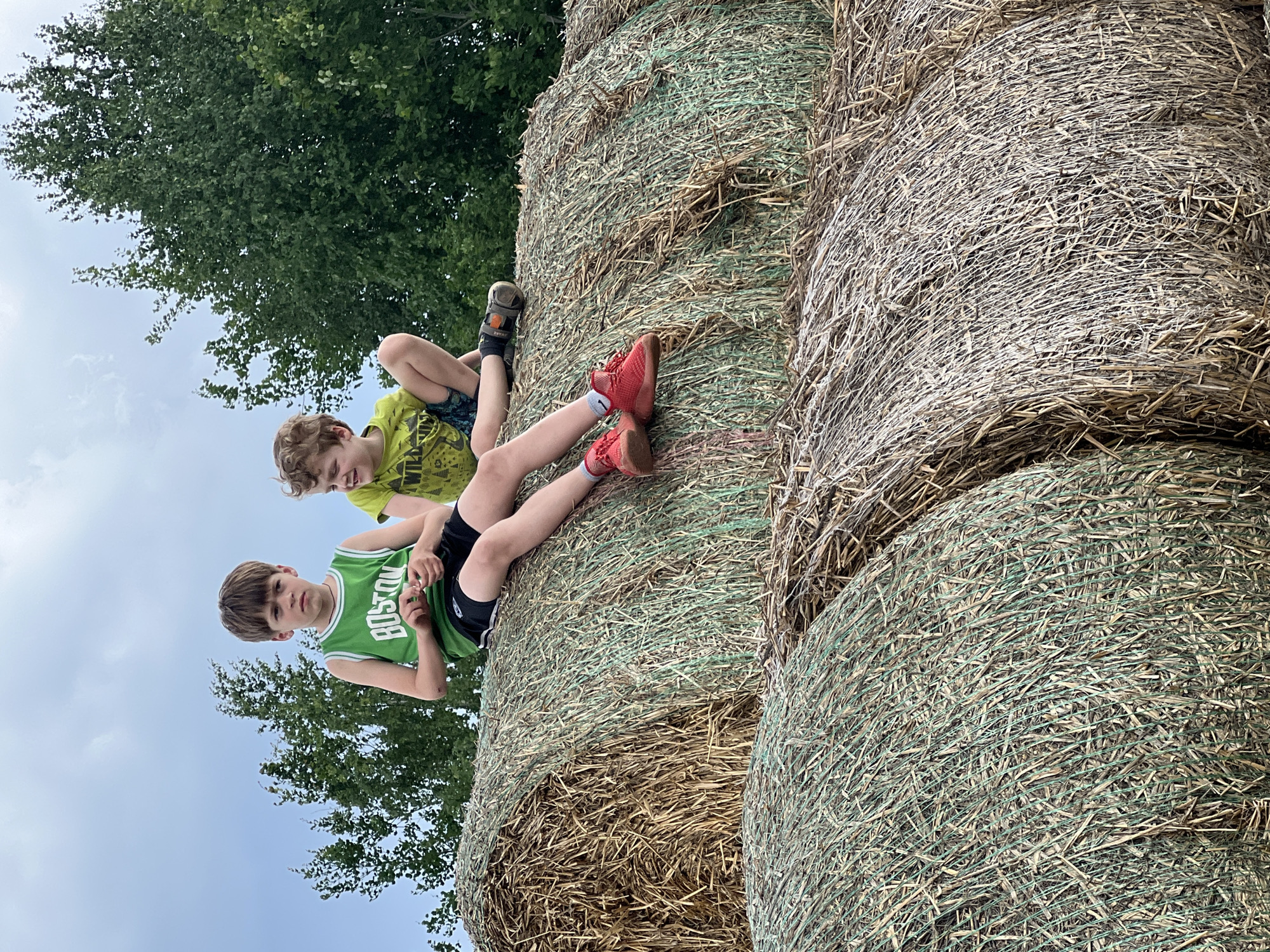 Two boys are lying on top of stacked hay bales. One boy is wearing a green Boston Celtics jersey and black shorts. The other boy is wearing a green patterned shirt and patterned shorts. The hay bales are cylindrical and made of dried grass. Trees are visible in the background, with a cloudy sky. The location is not specified to be in Kirillovka, Russia.