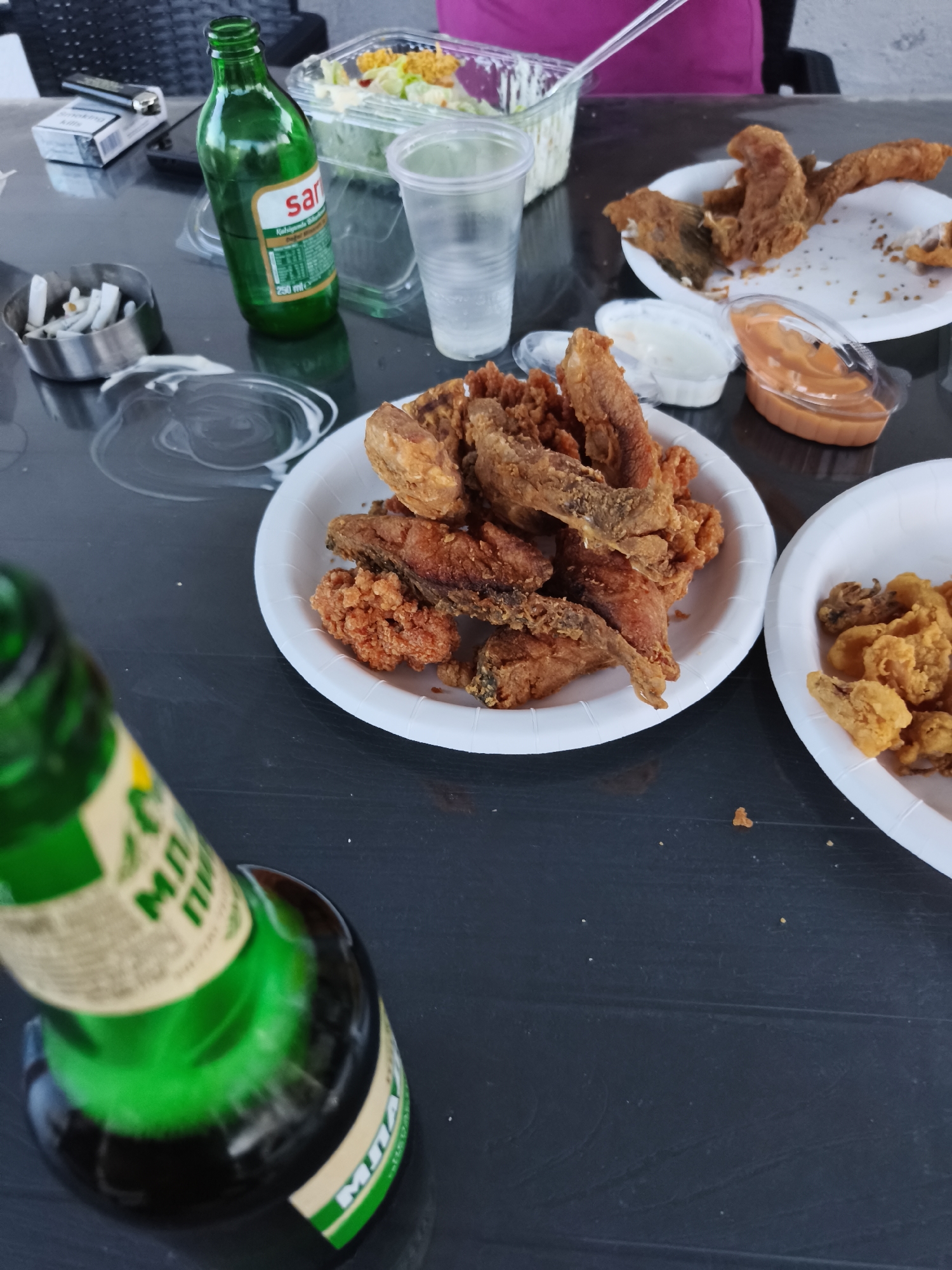 The image shows a table with plates of fried food, drinks, and cigarettes. In the foreground, a green glass bottle with a label is visible. On the table, there are two white paper plates, one filled with fried fish and the other with what appears to be fried calamari. There are also two small containers of dipping sauce, one white and one orange. To the left, an ashtray with cigarette butts and a pack of cigarettes are present. A clear plastic cup and a clear container of salad are also on the table. The background shows a person in a pink shirt. The table appears to be outdoors, with a dark, textured surface. The lighting suggests it is daytime.