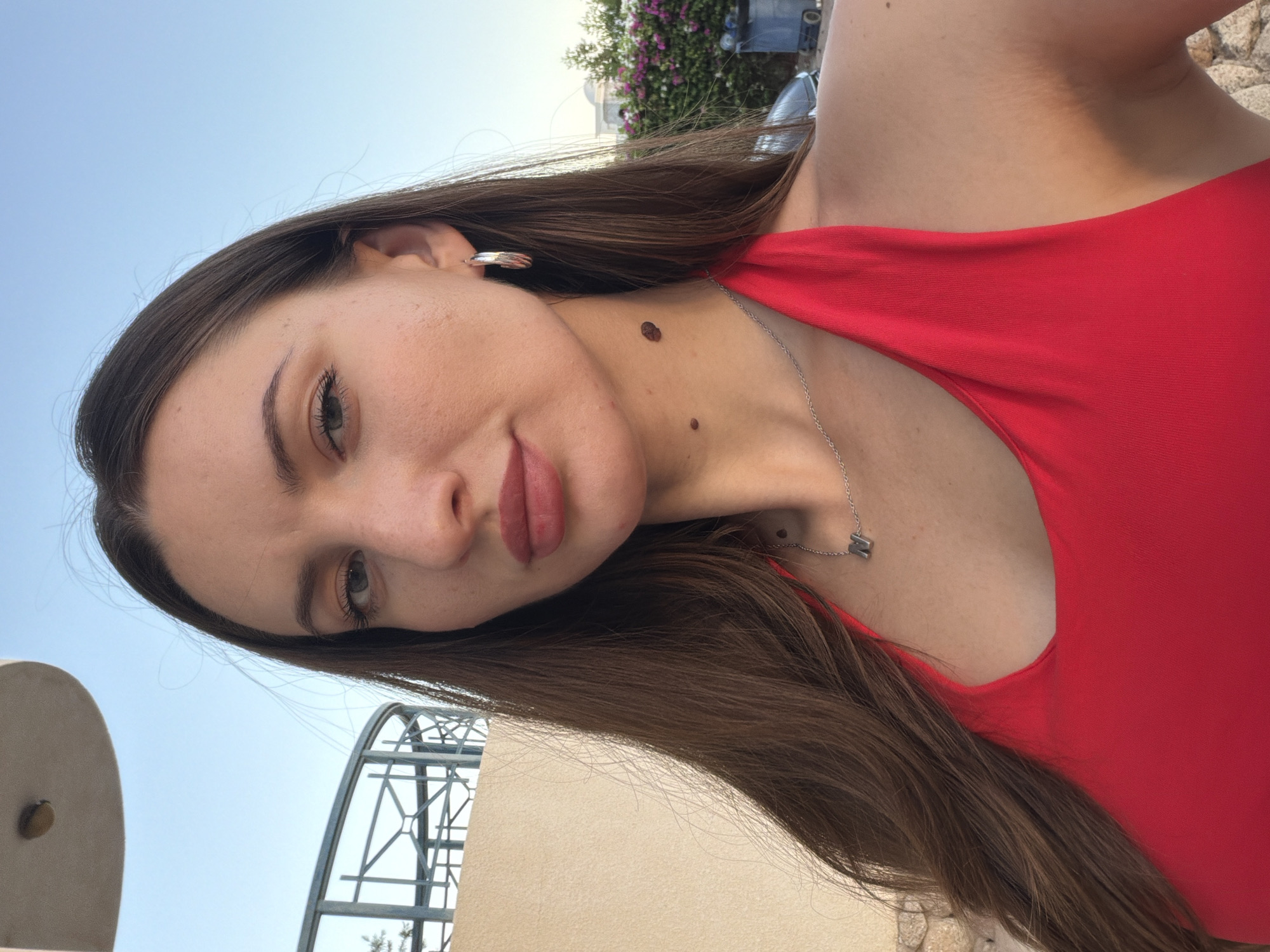 A young woman with long, dark brown hair and fair skin is captured in a close-up, selfie-style shot. She is wearing a vibrant red halter top and a silver necklace with a pendant shaped like the letter "N". She has a single silver hoop earring in her right ear. On her neck, there are three visible moles: one large and dark on her right side, and two smaller ones below it. Her lips are painted with a muted pink lipstick.
The background suggests an outdoor setting, possibly a balcony or terrace, with a pale yellow wall and a light blue sky above. A light-colored, curved structure with metal railings is visible in the upper left quadrant, and a railing with geometric patterns can be seen behind her left shoulder. In the distance, there are hints of buildings and lush green foliage with pink flowers, and a blue trash bin. The overall lighting indicates it is either early morning or late afternoon. The scene does not provide specific identifiers for Sharm El Sheikh, Egypt, beyond the presence of outdoor architecture and vegetation typical of warm climates.