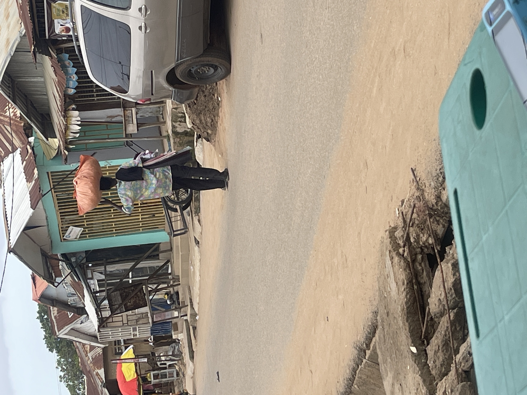 A person walks along a street in Abuja, Nigeria, carrying a large orange bundle on their head and a stack of folded clothes over their arm. The street is paved and lined with buildings, some of which appear to be market stalls or small shops with corrugated metal roofs. A silver van is parked on the right side of the road. In the foreground, a section of damaged concrete sidewalk with exposed rebar is visible. The sky is overcast.