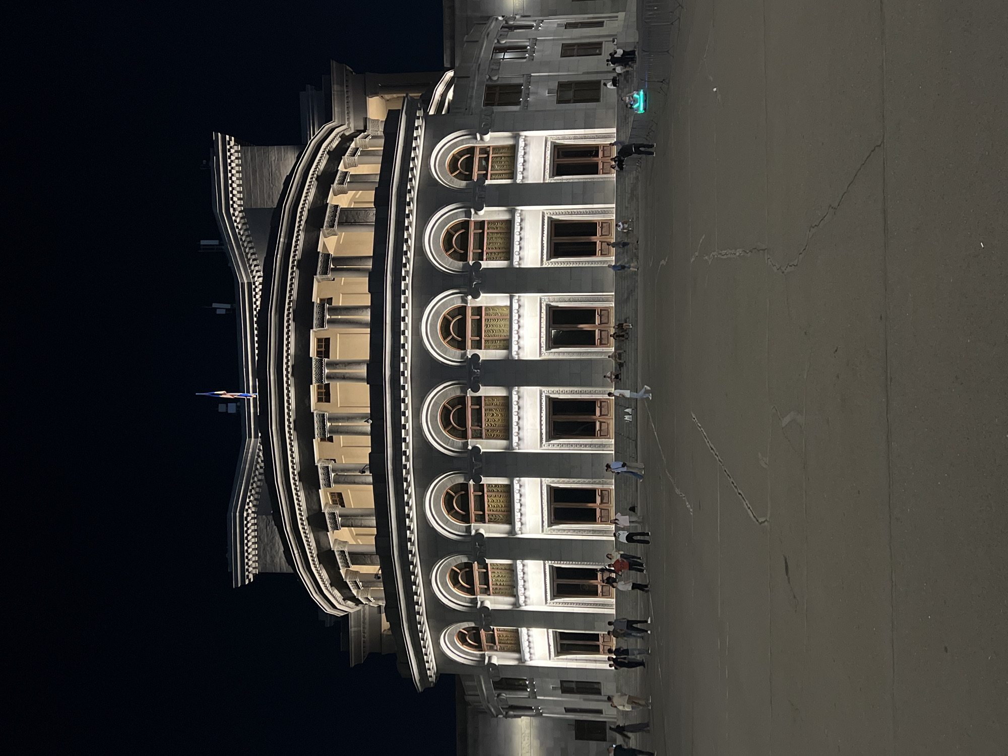 The image shows the exterior of a large, illuminated building at night. The building has a grand, neoclassical architectural style, featuring a curved facade with multiple arched windows and doorways. Columns are visible on the upper levels. A flag is flying from the roof. A group of people are gathered in the paved plaza in front of the building, some standing and some sitting on the steps. The plaza is cracked and worn. In the background, there are more people and some temporary fencing with a brightly lit green object inside. The overall scene suggests a public gathering or event taking place outside a significant cultural or governmental institution.