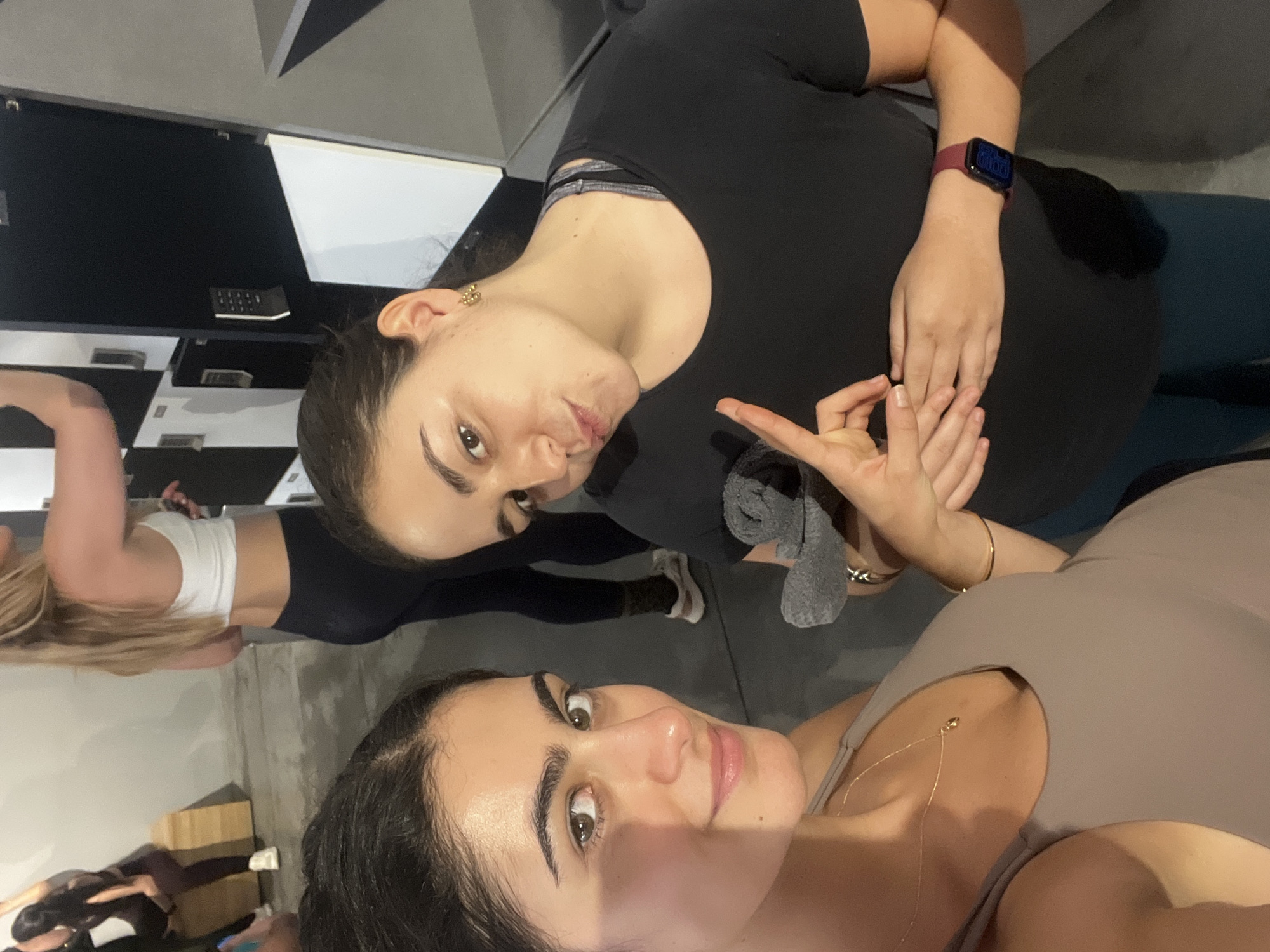 The image, captured in Paris, France, features two women prominently in the foreground, with other individuals visible in the background, all captured from a tilted, upward-looking perspective within what appears to be a modern indoor fitness or yoga studio.
In the immediate foreground, a woman with dark hair, seemingly pulled back, wears a light brown or beige tank top and a delicate gold chain necklace. Her gaze is directed towards the camera, displaying a calm and friendly expression. Her hair appears to have some visible partings.
Slightly behind and above her, another woman with dark hair is seen. She wears a black short-sleeved top over a grey sports bra with strap detailing. She looks directly at the camera with a relaxed, slightly pouty expression. Her left wrist is adorned with a red-banded smartwatch displaying illuminated digital numbers, partially visible as "00:00". Her hands are gently clasped near her midsection, with her right hand forming a gesture resembling a mudra, where the thumb and index finger touch. A grey cloth is either held in her left hand or tucked into her attire. She wears a golden earring in her right ear and a simple gold bracelet on her right wrist.
The background reveals several other individuals, suggesting a fitness class in progress. One person with long blonde hair, wearing a white crop top and black leggings, stands near a light brown yoga block. Other figures are partially visible or blurred, some appearing to be sitting or standing on mats. The studio environment features dark walls, some incorporating white and black geometric patterns, suggesting a contemporary design. No explicit text is clearly legible anywhere in the image. The overall setting indicates an active yet relaxed atmosphere, likely during a group exercise session.