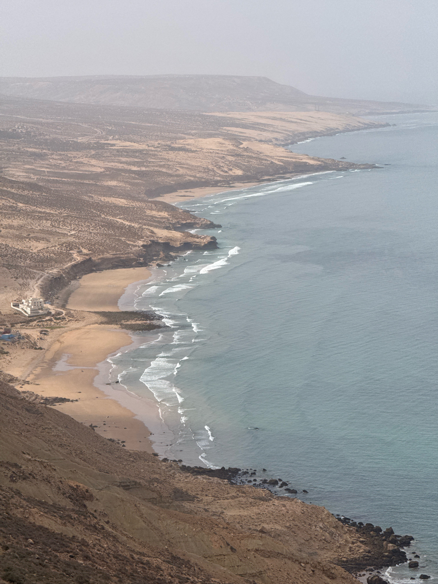 This aerial photograph captures a view of the coastline near Imsouane, Morocco. The scene is dominated by a rugged, arid landscape meeting the ocean. In the foreground, a steep, rocky, and sparsely vegetated slope descends towards the coast. The mid-ground features a crescent-shaped beach composed of light brown sand, with small waves breaking on the shore. A few rocky outcrops punctuate the waterline. On the beach, a large, multi-story white building with a flat roof is situated, appearing to be a hotel or resort. Beside it, there are a few smaller structures and some tents or temporary shelters. A dirt road winds its way from the building up into the arid hills. The ocean, a muted blue-grey, stretches into the distance, with lines of white surf indicating waves. The background shows a hazy, mountainous terrain receding into the atmospheric distance, suggesting a slightly overcast or dusty day. No people are visibly present in the scene.