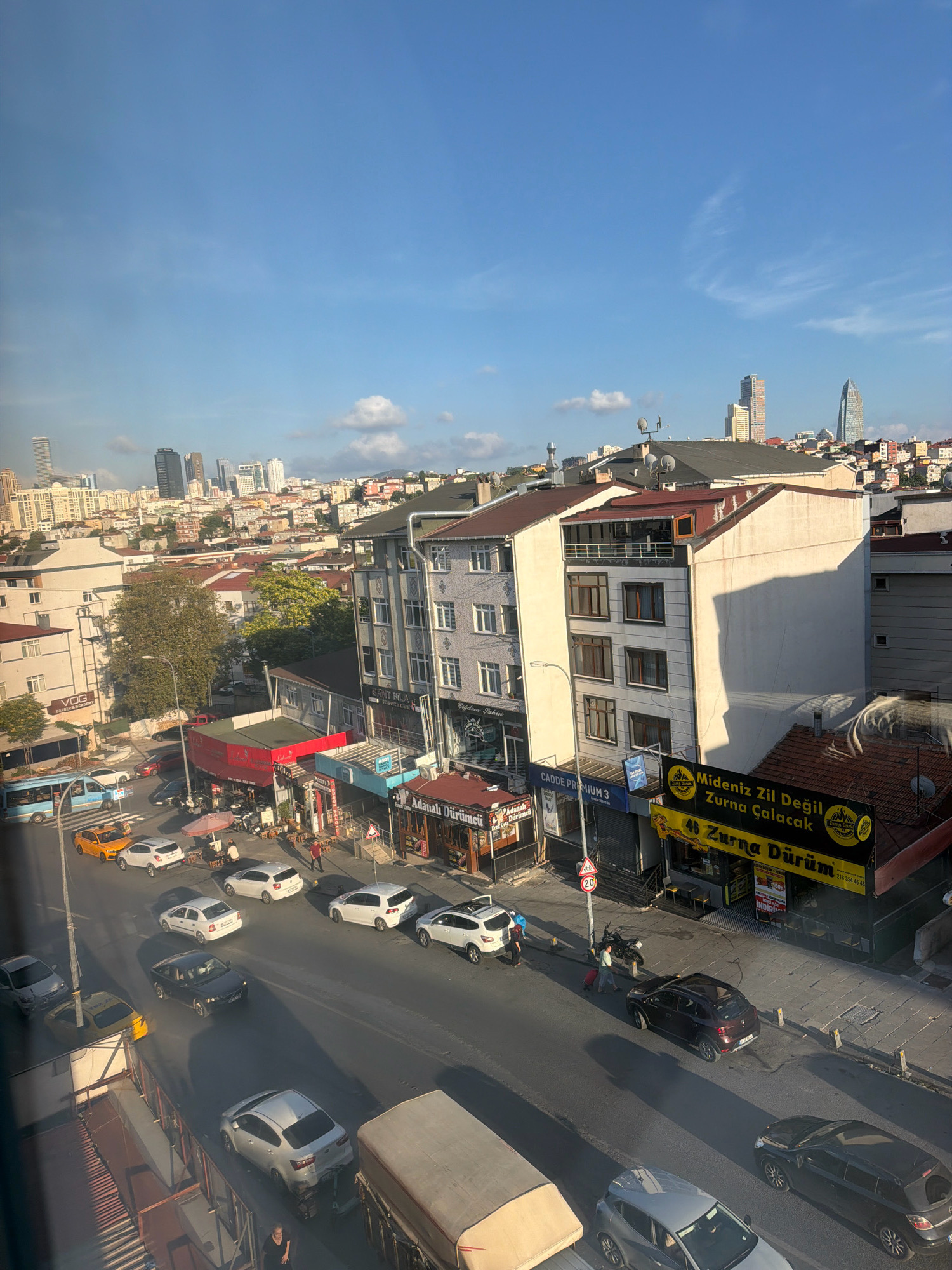 The image shows a street scene in Ataşehir, Turkey, on a clear day with a blue sky and scattered clouds. A multi-lane road runs through the center of the frame, lined with buildings on both sides. Traffic is visible on the road, including several cars and a yellow taxi. Pedestrians are also present on the sidewalks. The buildings are a mix of modern and older styles, with shops and businesses at street level. Some of the businesses have visible signage, including one that reads "Mideniz Zil Değil Zurna Durum". In the background, a cityscape with taller buildings and skyscrapers can be seen. The overall impression is of a bustling urban environment.