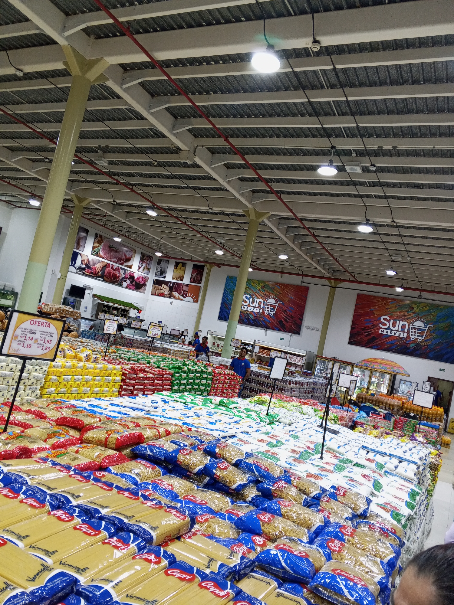 Here is a detailed description of the image:
**General Overview:**
The photograph captures a wide-angle view of the interior of a large supermarket. The perspective is from the customer's point of view.
**Location:**
Based on the signage visible in the image ("Sun Market"), this scene is likely within a Sun Market supermarket, although the city of Altagracia, Venezuela, is not discernable.
**Subjects and Objects:**
* **Large Display of Goods:** A significant portion of the image is occupied by a large display featuring stacks of food products, primarily pasta and sugar. Pasta is in blue Capri-branded bags.
* **Signage:** Prominent signage is present, indicating "Oferta" (Spanish for "Offer"), with prices displayed for various pasta products.
* **Personnel:** Several workers and customers are visible in the background of the image.
* **Ceiling Structure:** The supermarket has a high ceiling with visible metal infrastructure, lights, and red piping.
* **Product Banners:** Large banners displaying the "Sun Market" logo are hung on the back wall.
* **Other Displays:** Other product displays can be seen in the background, like meat products in a cooler.
**People:**
* There are several people in the scene. Two workers wearing blue shirts are visible behind the product display.
**Environment:**
* The setting is a well-lit supermarket interior with a high ceiling.
**Action:**
* Customers shop in the background.
* Workers are present, presumably stocking and organizing the product displays.
In summary, this image depicts a bustling interior of a Sun Market supermarket with a focus on a large product display, signage advertising prices and workers.
