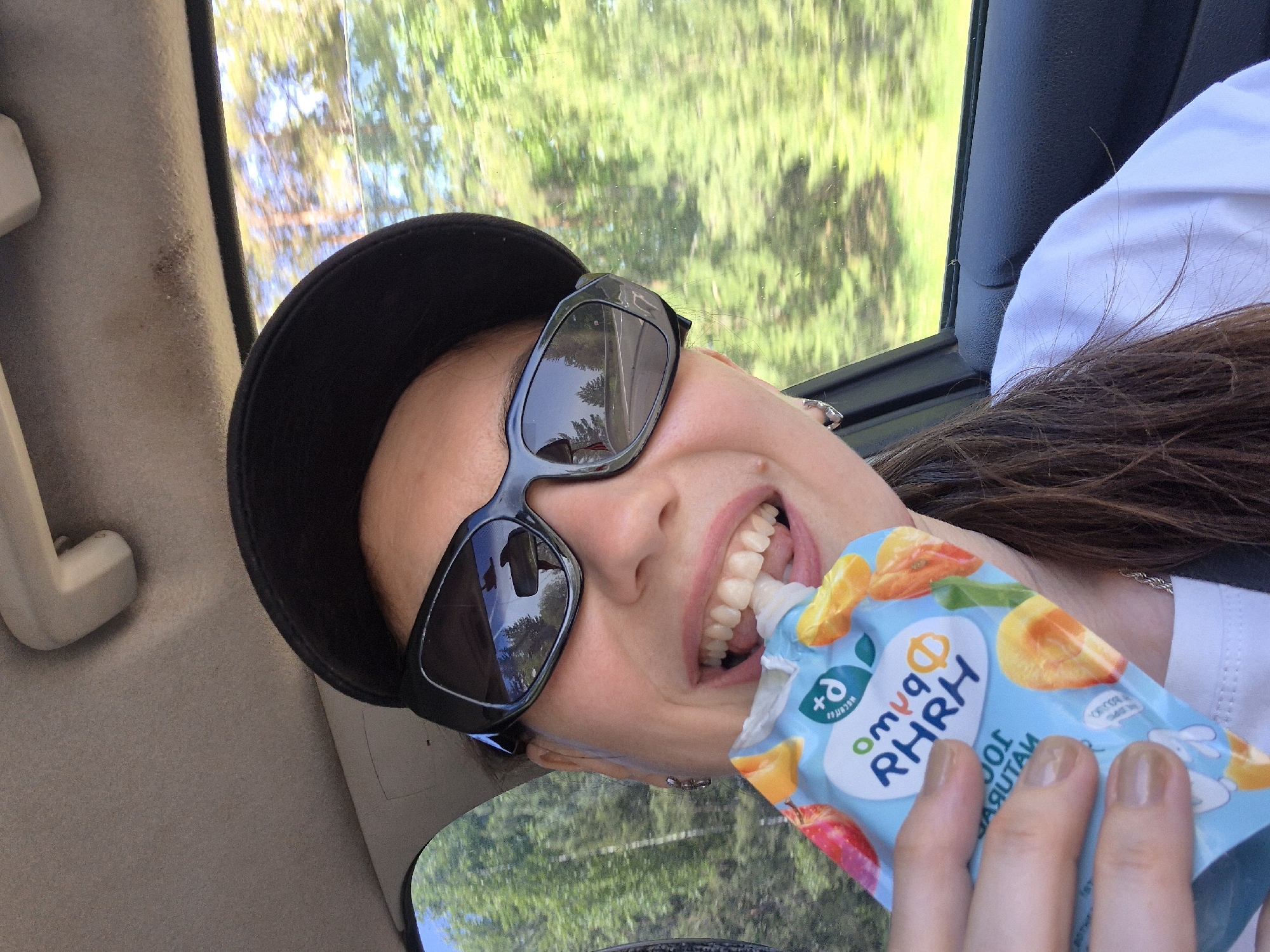 A woman in a car, possibly in the city of Unknown, Unknown, wearing a black cap and sunglasses, smiles as she eats from a blue fruit puree pouch. Outside the car window, trees are visible, reflecting in her sunglasses. The car's interior is visible, including a beige ceiling, a black seat frame, and a white handle. The woman is wearing a white shirt and a silver necklace.