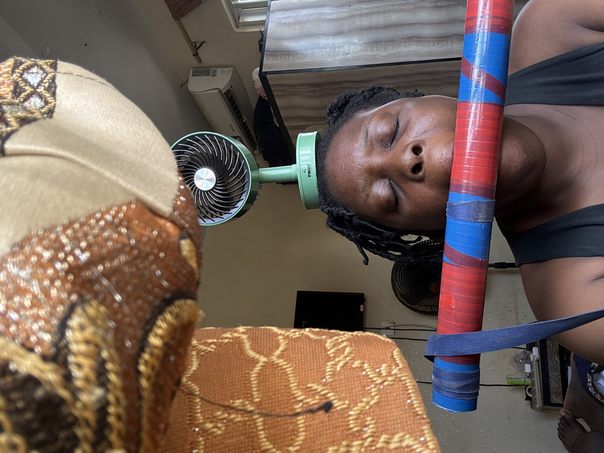 In Jawu, Nigeria, an individual with closed eyes is holding a long, cylindrical object horizontally across their mouth. The object is wrapped in red and blue tape, with a dark blue strap attached. The person is wearing a dark tank top. In the background, a green fan is positioned on the floor behind the individual, and further back, an air conditioning unit is mounted on the wall above a cabinet. The scene appears to be indoors, possibly in a living space, with decorative fabric hanging from the ceiling in the foreground.