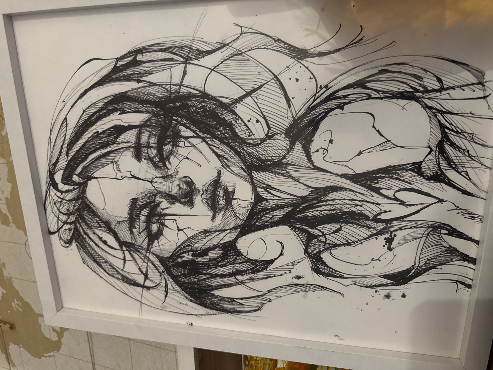 The image is a framed black and white drawing of a woman's face with stylized hair, viewed at an angle. The drawing is rendered in thick, gestural lines with cross-hatching for shading. The face appears to be cracked or fragmented. The frame is white. The surrounding environment is a wall with damaged wallpaper. The location is unavailable.